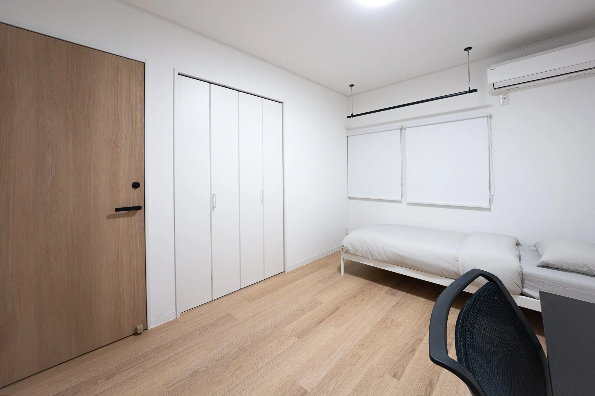 A minimalist bedroom with light wooden flooring, a white bed positioned against the right wall with two pillows, white window blinds above the bed, a large wooden door on the left, and a white wardrobe with three doors. There is an air conditioning u