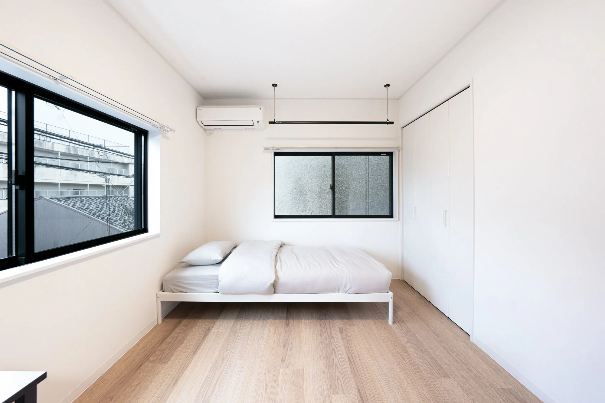 Minimalist bedroom with white walls, wooden floor, a single bed with white bedding, two windows, a white closet with sliding doors, and a wall-mounted air conditioner.