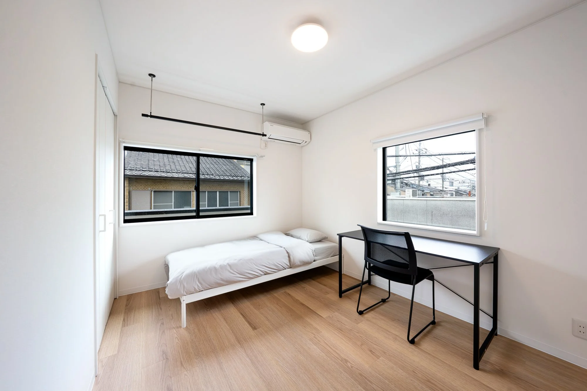 A minimalist bedroom with white walls, two black-framed windows, a single bed with white bedding, a black desk with a black chair, and wooden flooring.