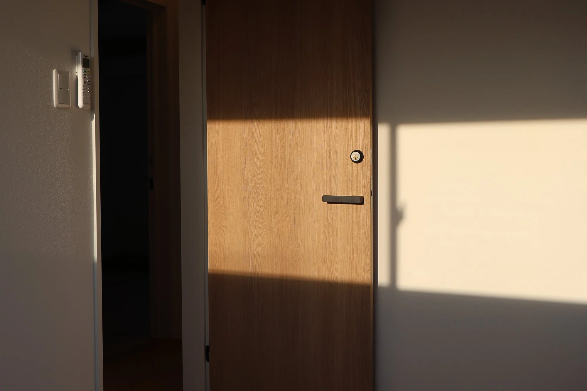 A wooden door partially open, with sunlight casting a shadow on a white wall beside it.