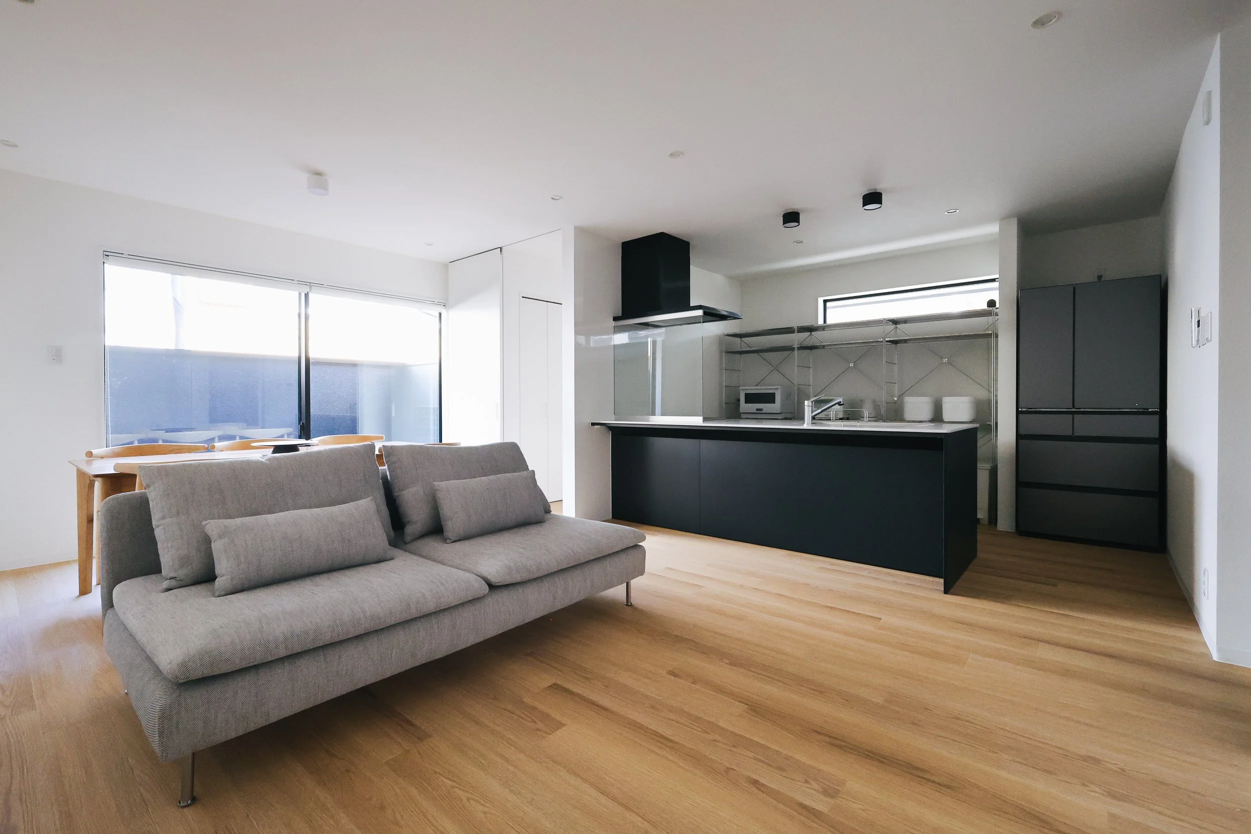 Modern open-concept living area with a gray sofa, light wood flooring, large windows, and a kitchen with dark lower cabinets, a black range hood, and shelves with white dishes.