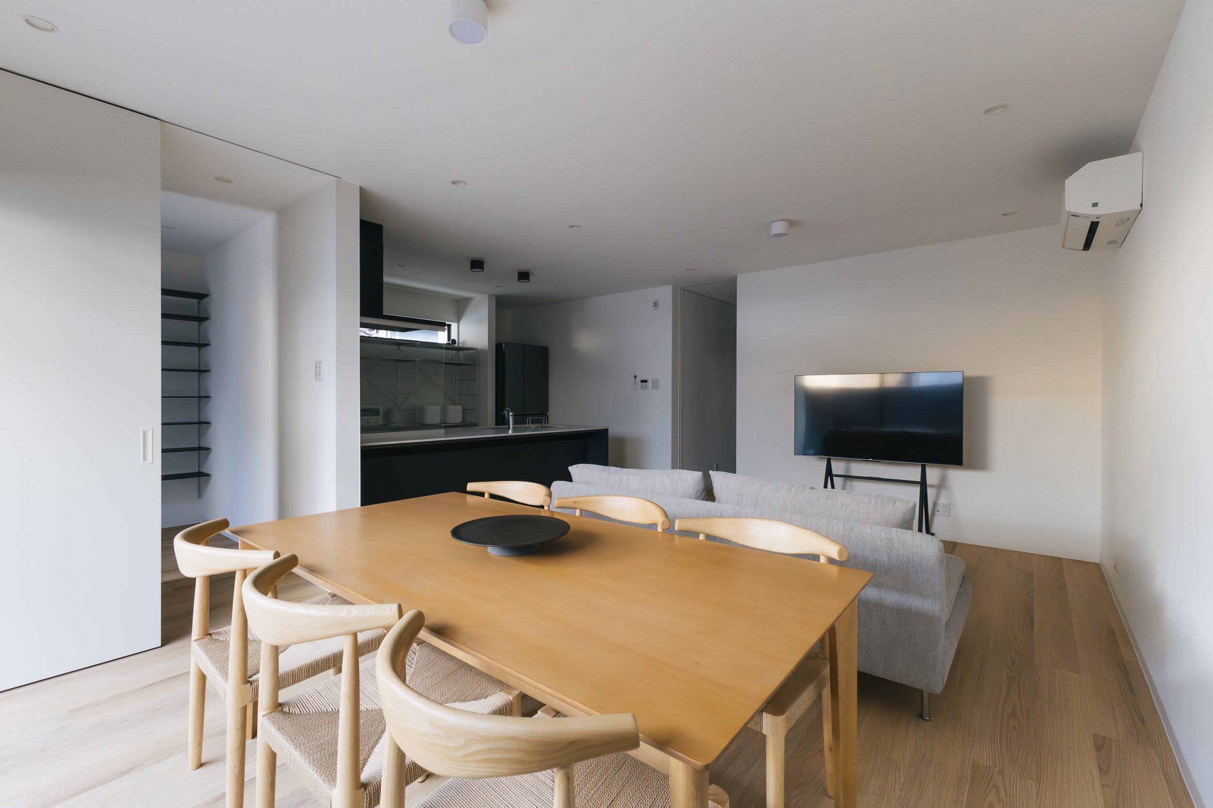 A modern, open-concept living room with a dining area that includes a wooden table surrounded by light wood chairs with woven seats. In the background, there's a kitchen with dark cabinets, a stainless steel refrigerator, and a window, along with a w