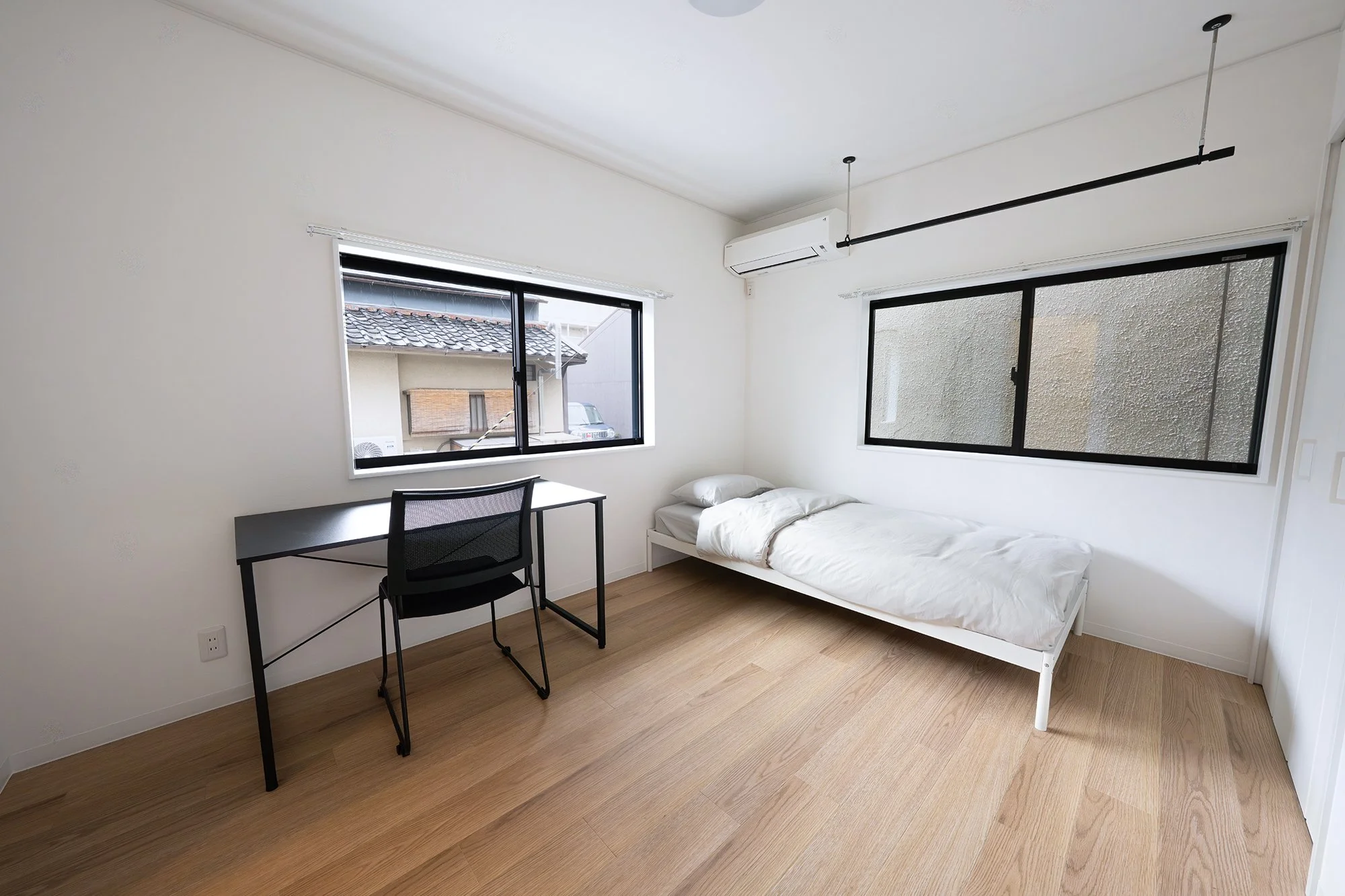 Minimalist bedroom with a single bed, pillows, white bedding, black-framed windows, a black desk, and a black mesh chair. Light wooden floor and white walls.