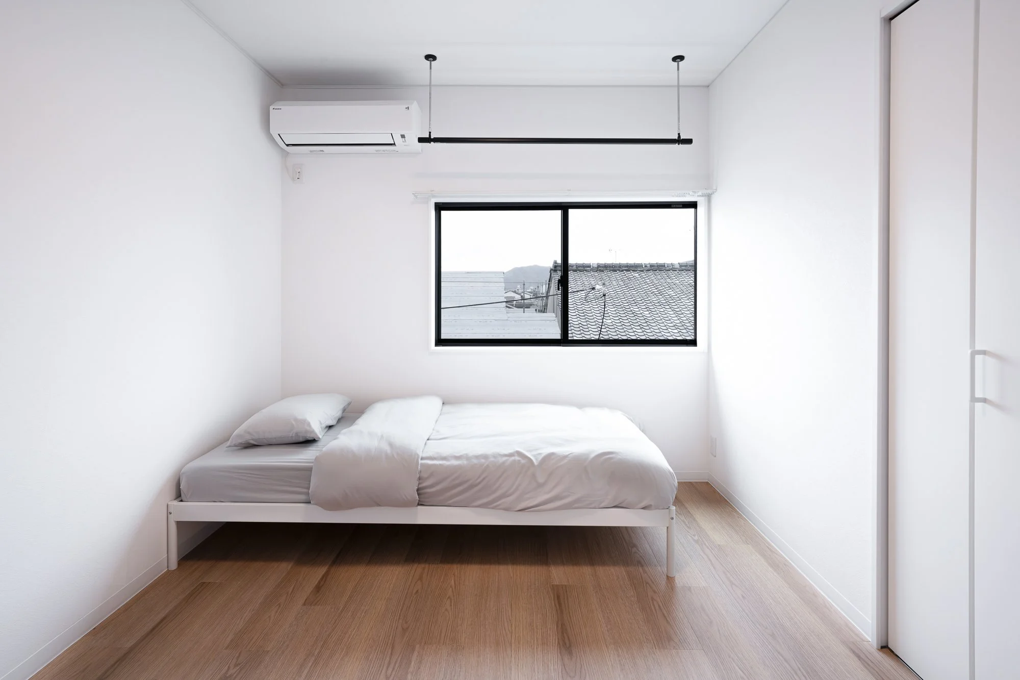 Minimalist bedroom with white walls, a bed with white bedding, two pillows, a large window, and wooden flooring.