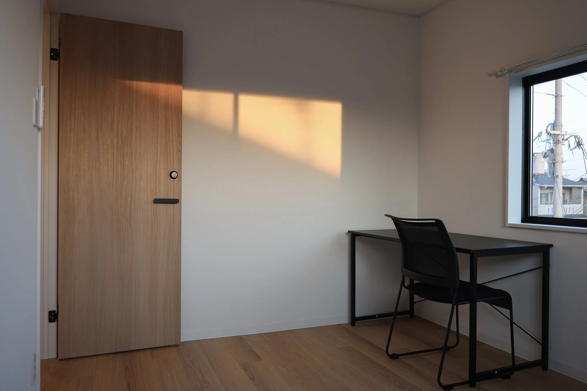 Empty room with a wooden door on the left, a black desk and chair near a window on the right, sunlight casting a rectangular shadow on the wall.