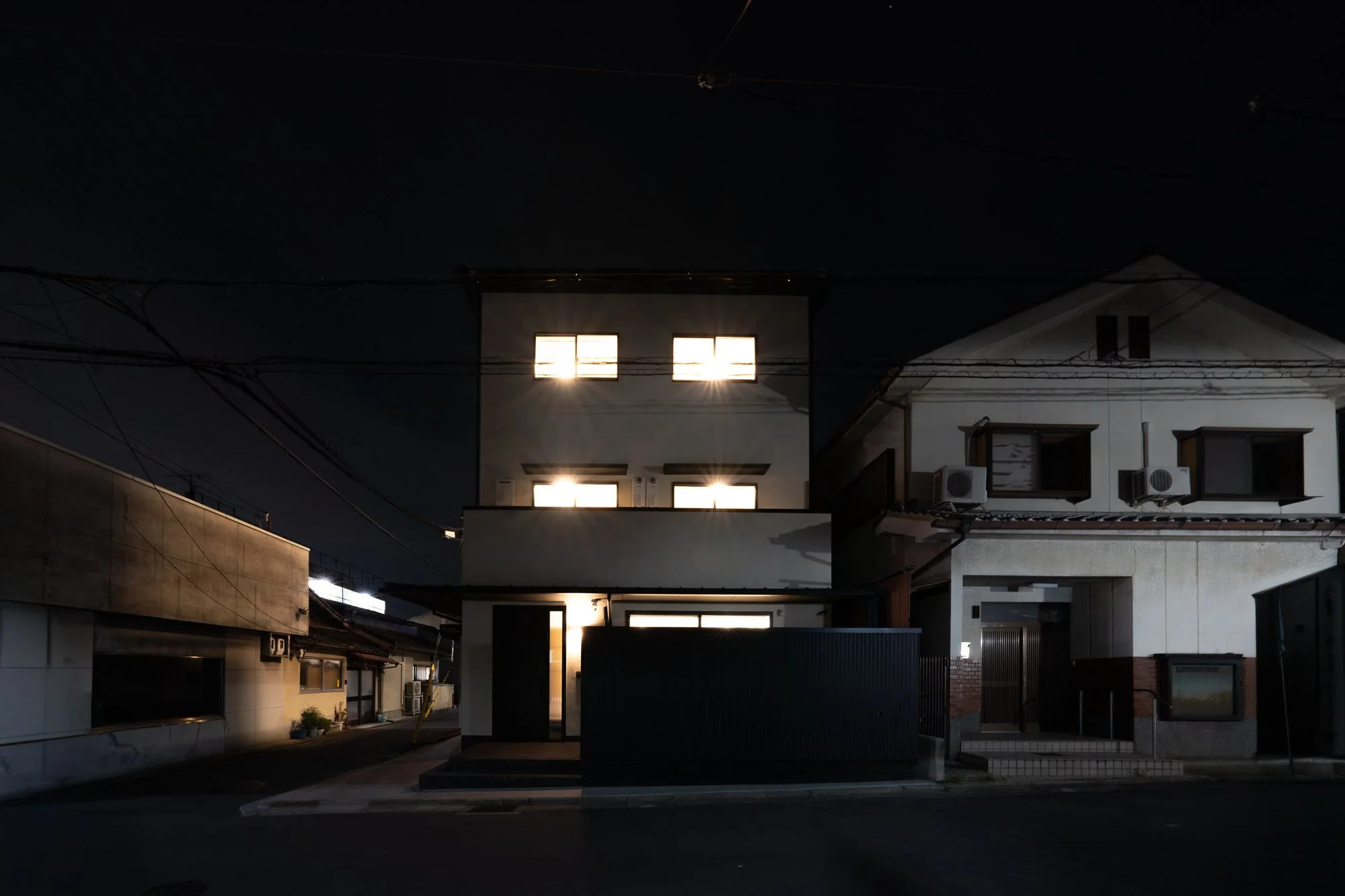 Night view of a modern house with illuminated windows and neighboring buildings, dark sky, and power lines.