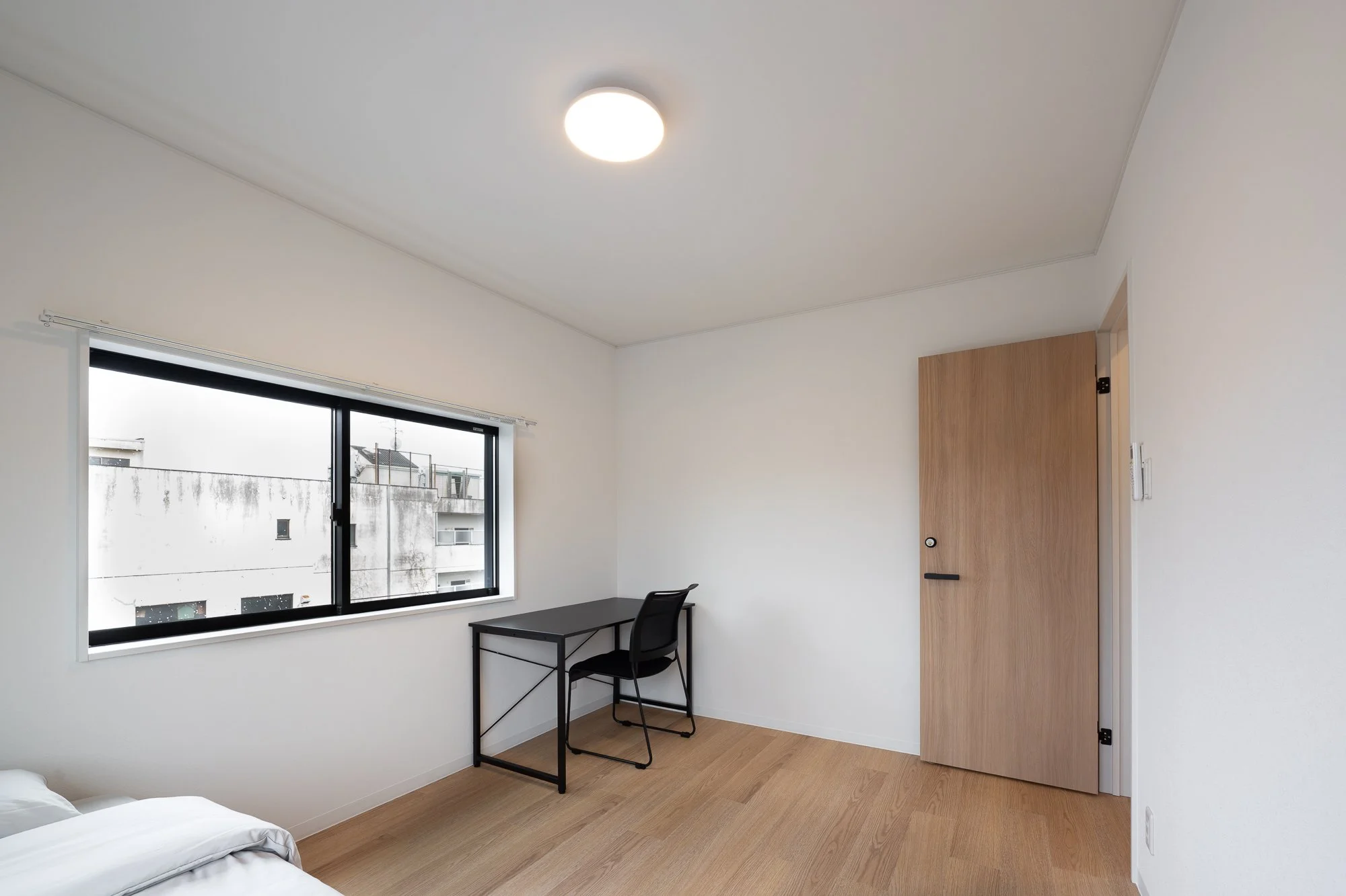 Empty room with a window, a black desk with a chair, a wooden door, and a ceiling light.