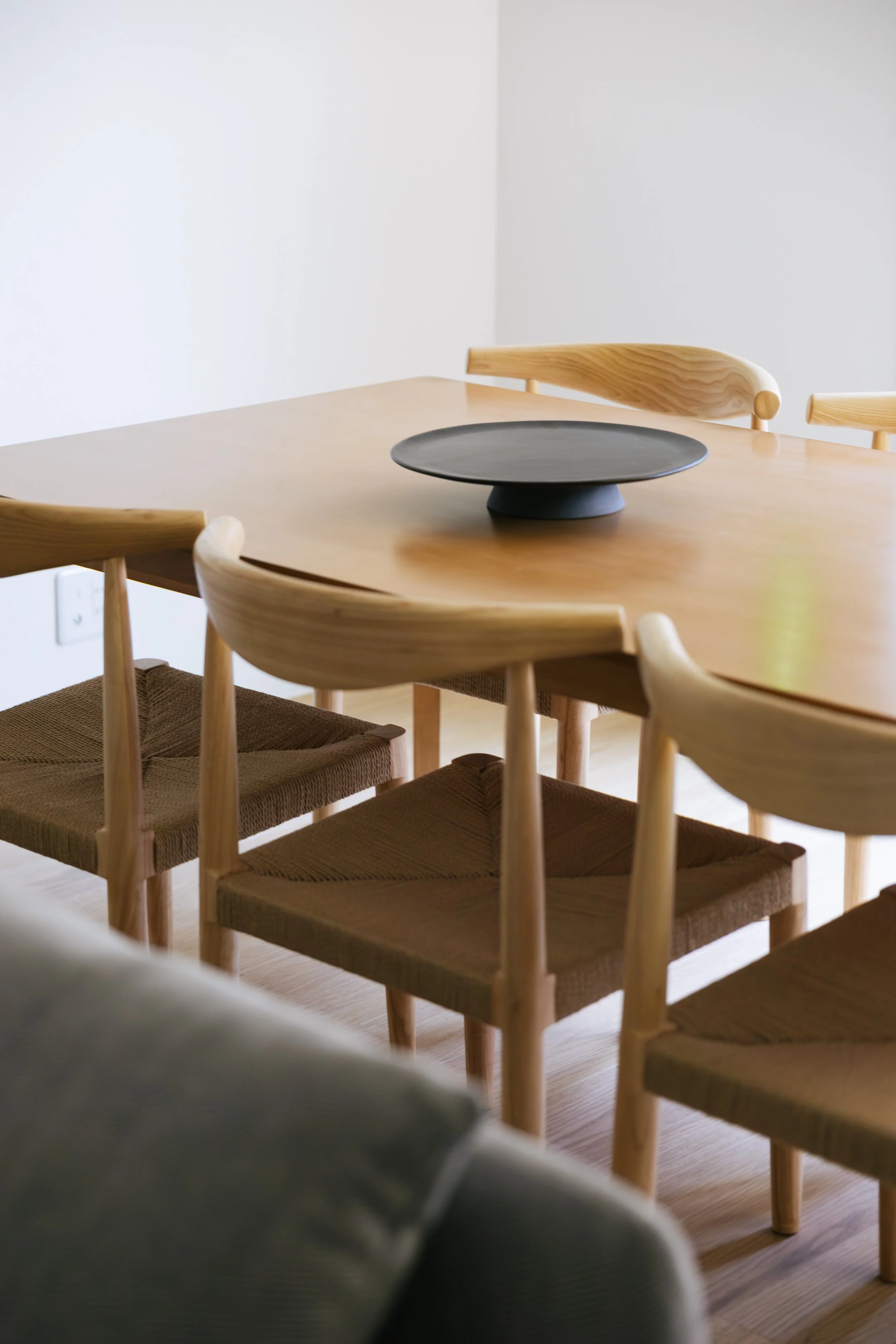 A wooden dining table with four wooden chairs and a black decorative tray in the center, in a minimalist room with white walls and light wood flooring.