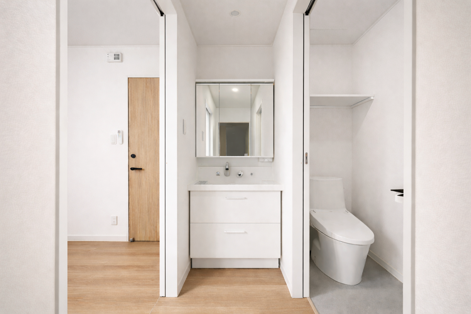 A small bathroom with white walls and a wooden door, featuring a white vanity with a mirror, a sink, and a modern toilet situated in a separate enclosed space.