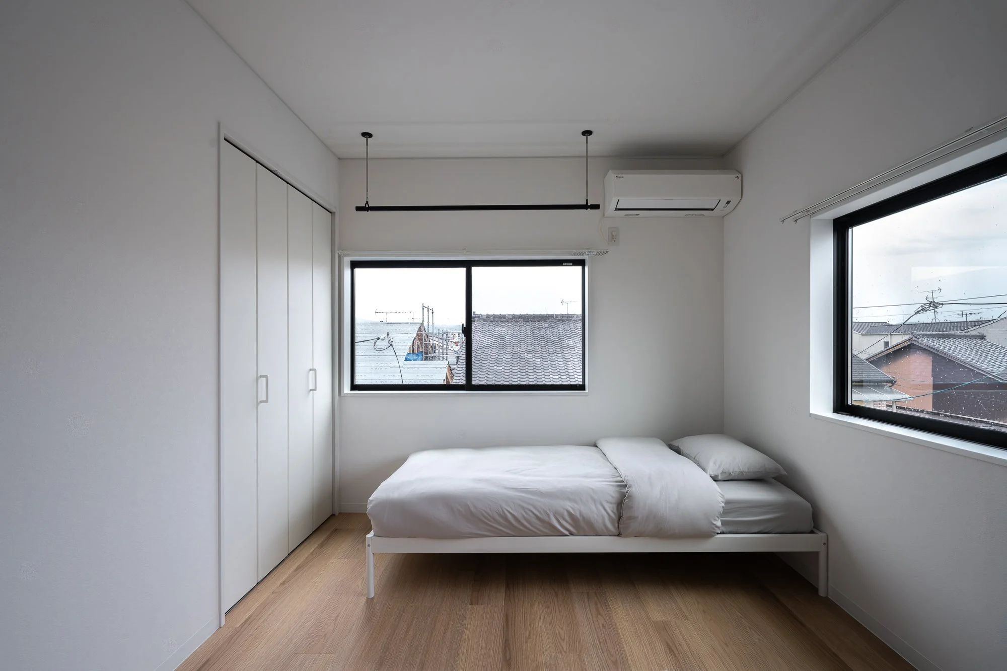 Empty bedroom with white bed, two windows, white closet, and wooden floor.