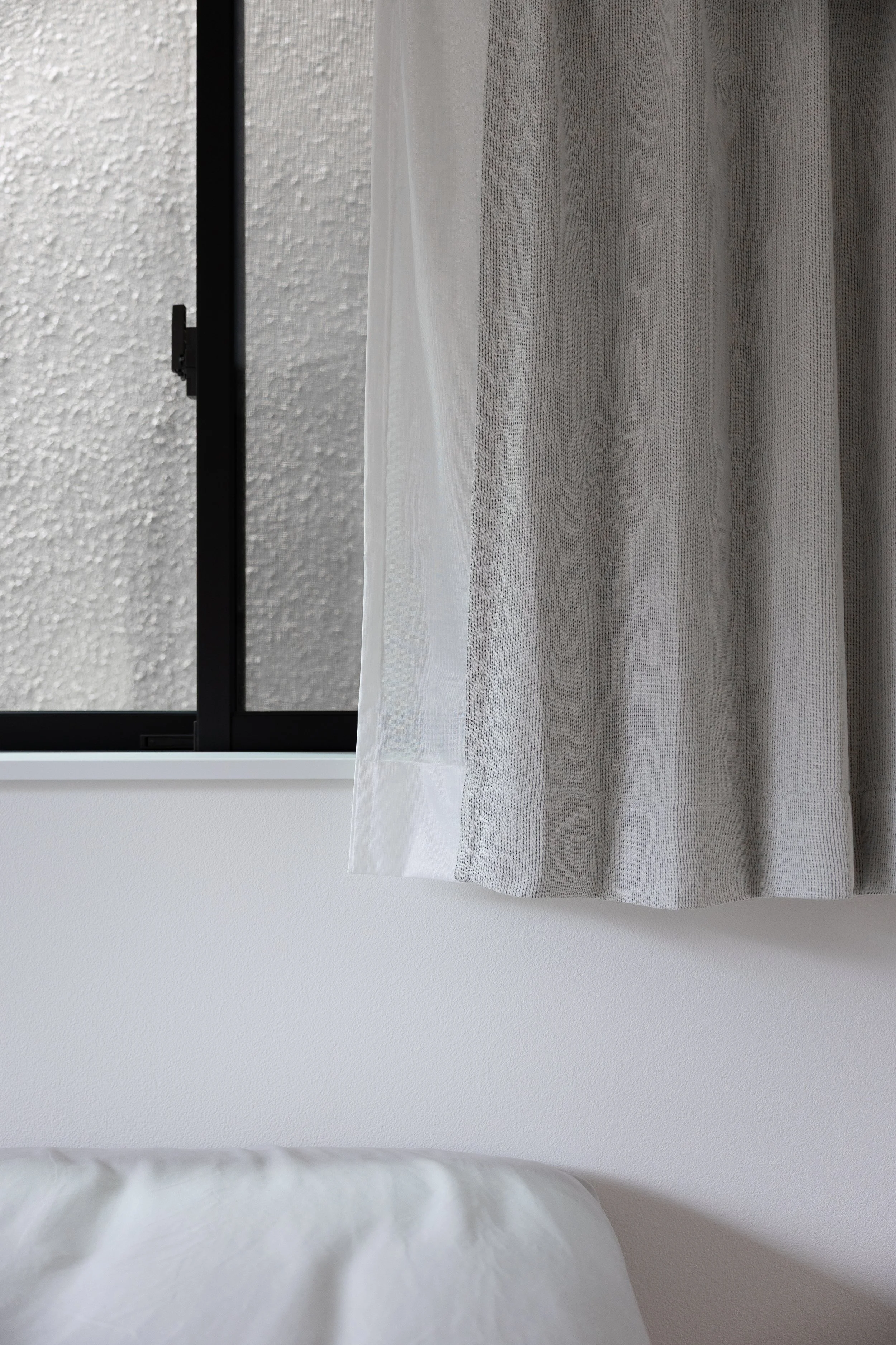 Close-up of a window with frosted glass, black frame, and a sheer white curtain partially covering the glass, with a white wall underneath.