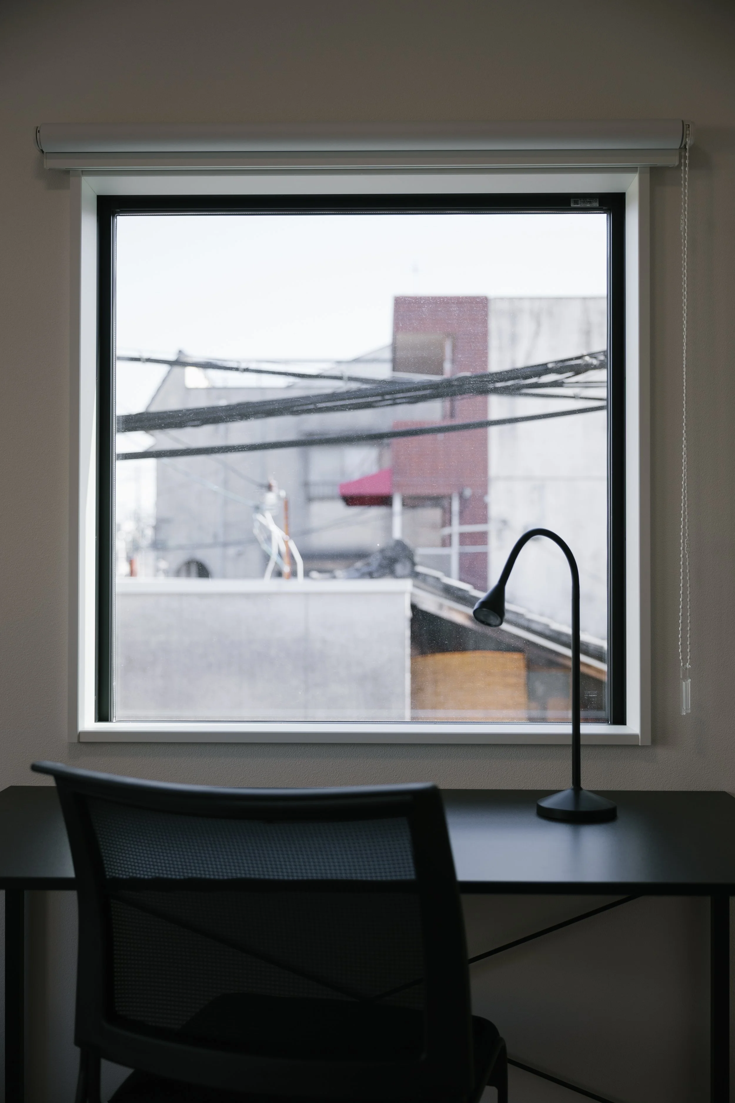 An office interior featuring a black mesh office chair, a desk, a window with a roller blind, and a black desk lamp. The window provides a view of neighboring buildings and power lines.