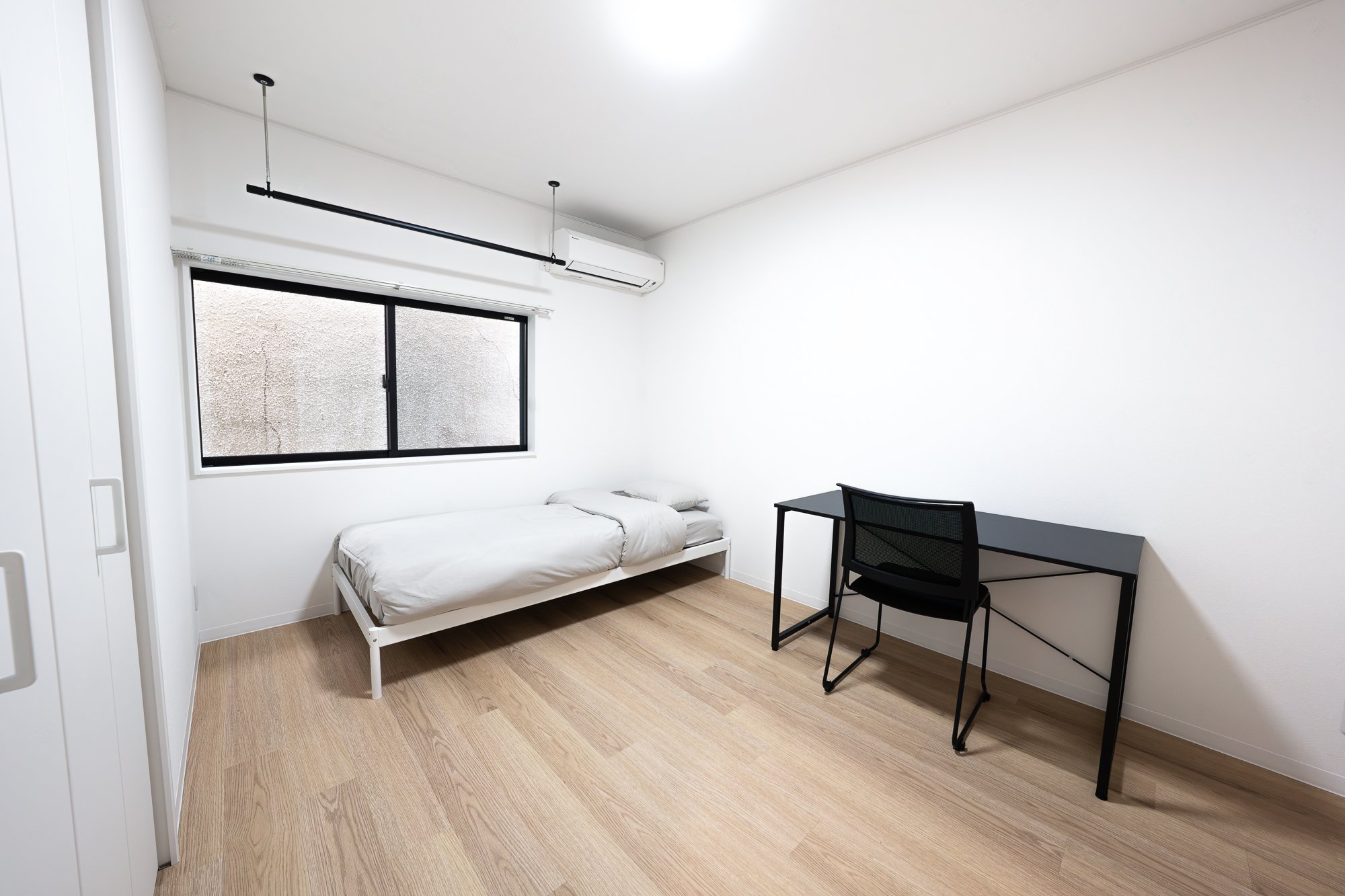A minimalistic bedroom with a single bed, a desk, a black chair, and a window with a view of a concrete wall.