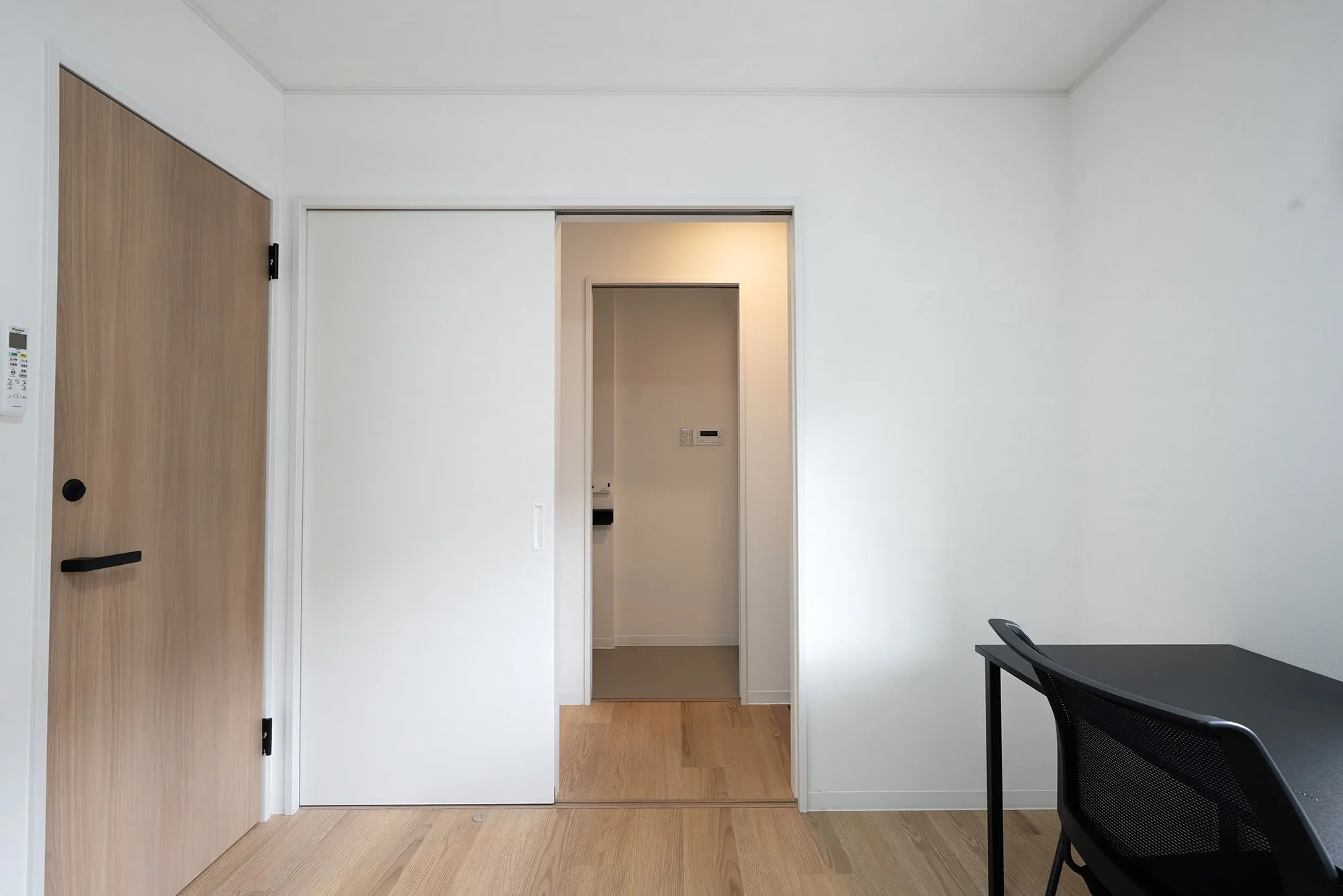 An empty room with white walls and light wood flooring, featuring a closed wooden door on the left and an open sliding door leading to another room or corridor, with a black office chair and a black table on the right side.