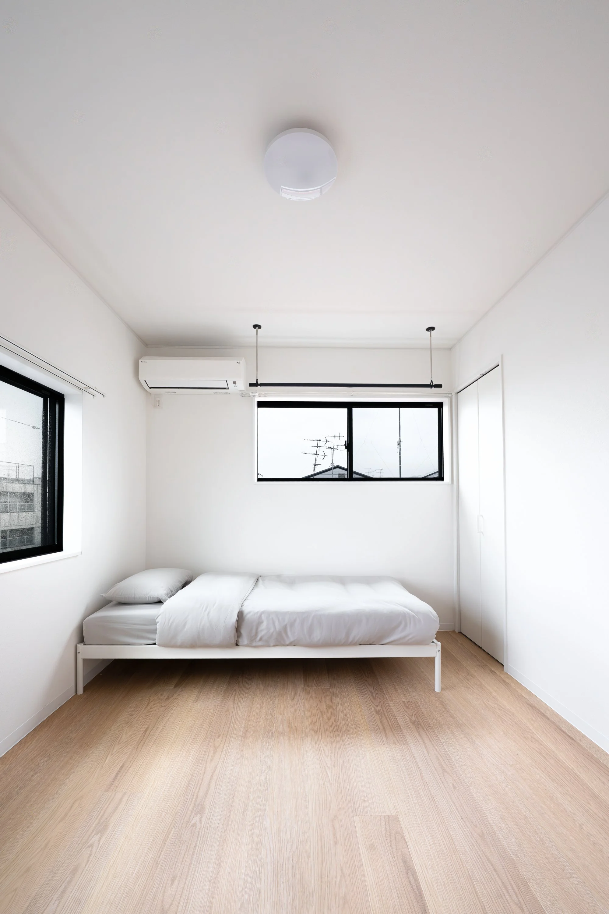 Empty white bedroom with a single bed, two windows, an air conditioner, a ceiling light, and a closet door.