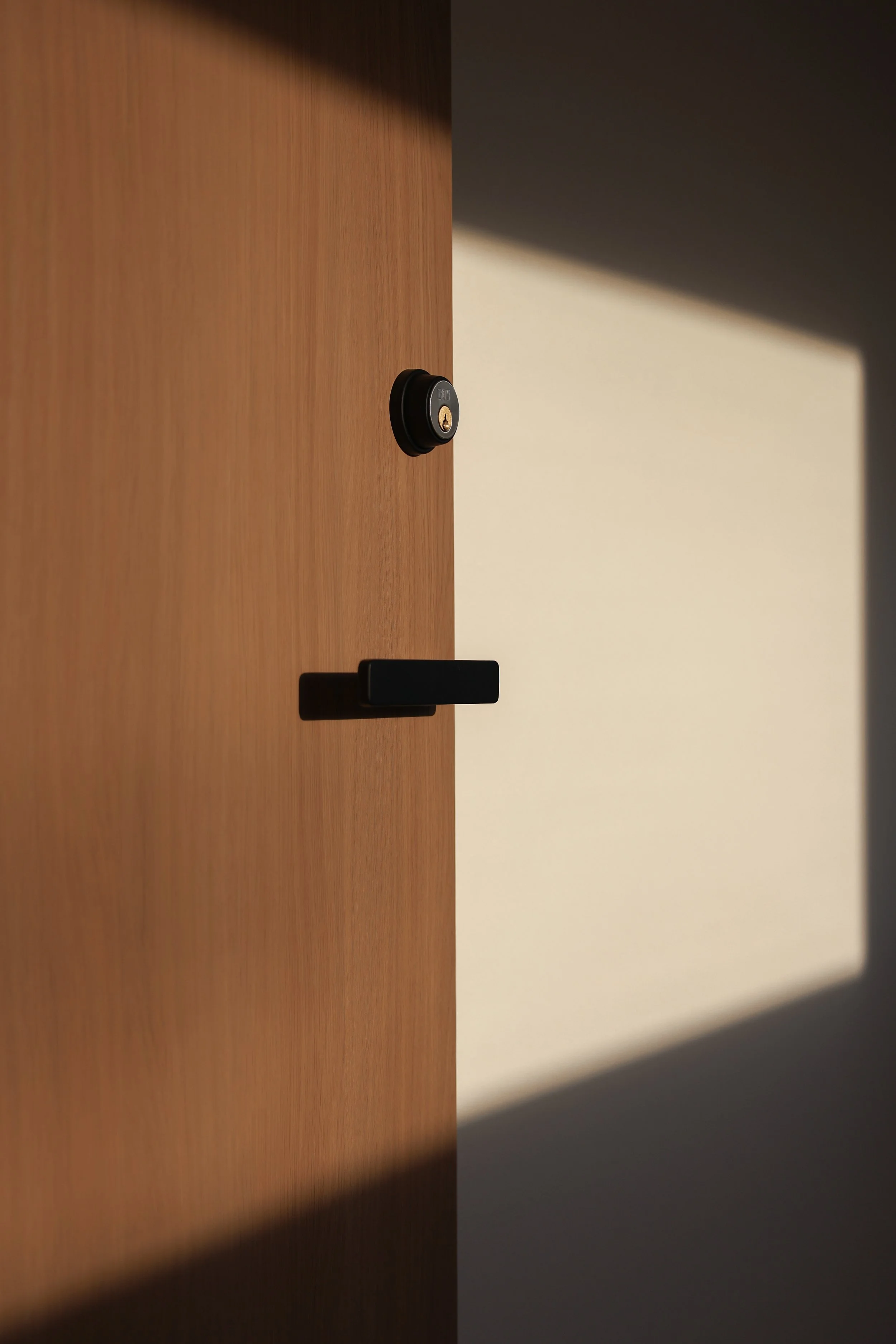Close-up of a wooden door with a black electronic lock and a black handle, with sunlight casting a shadow of the handle on a white wall.