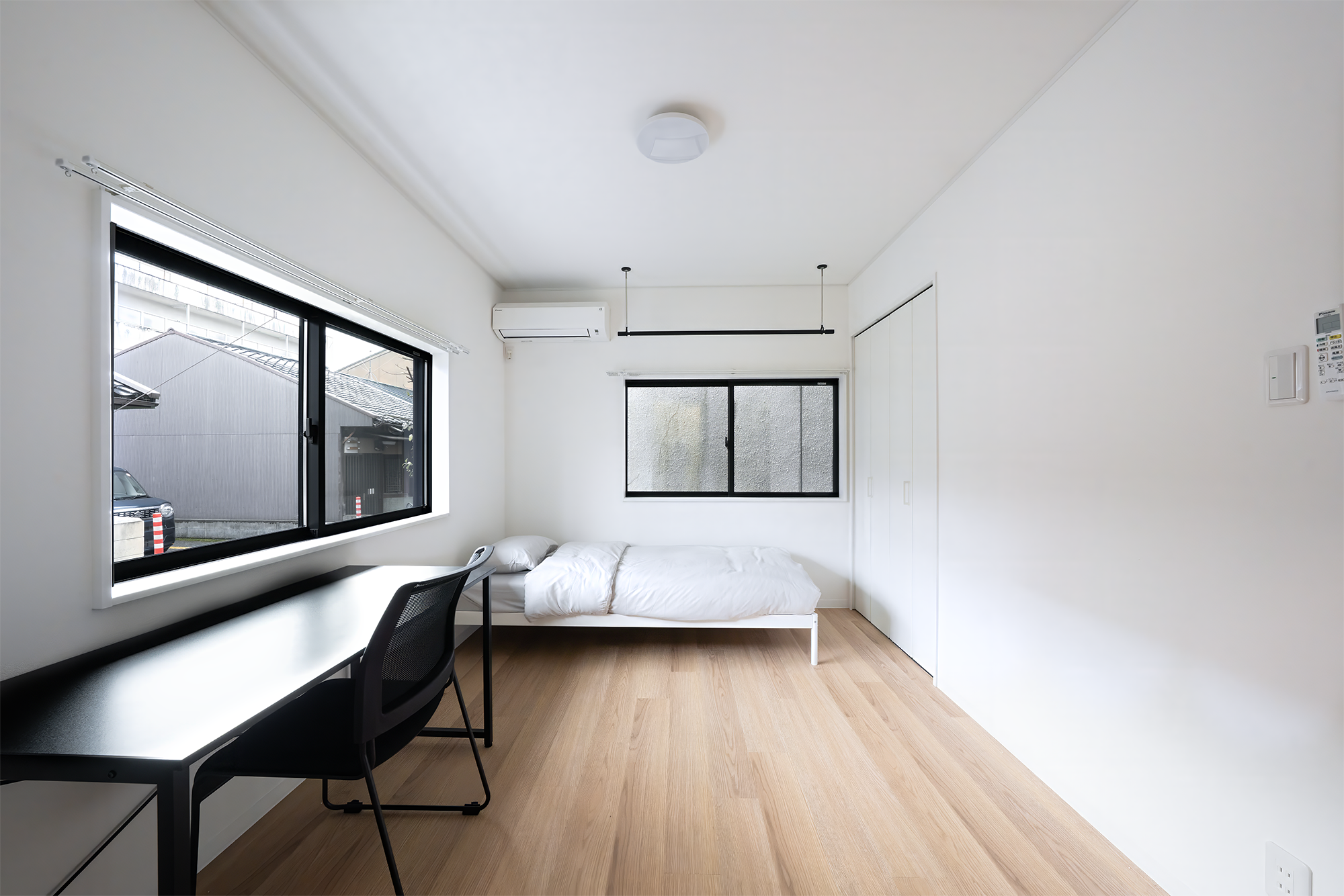 Minimalist bedroom with a bed, black desk, and chair, with two windows and wooden floors.