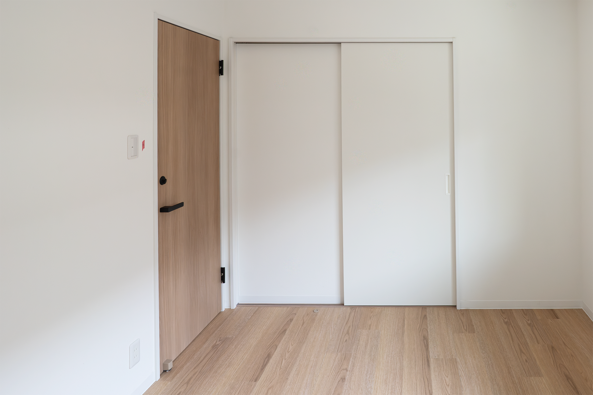 Empty room with white walls, a wooden door with black handle, sliding white closet doors, and light wood flooring.