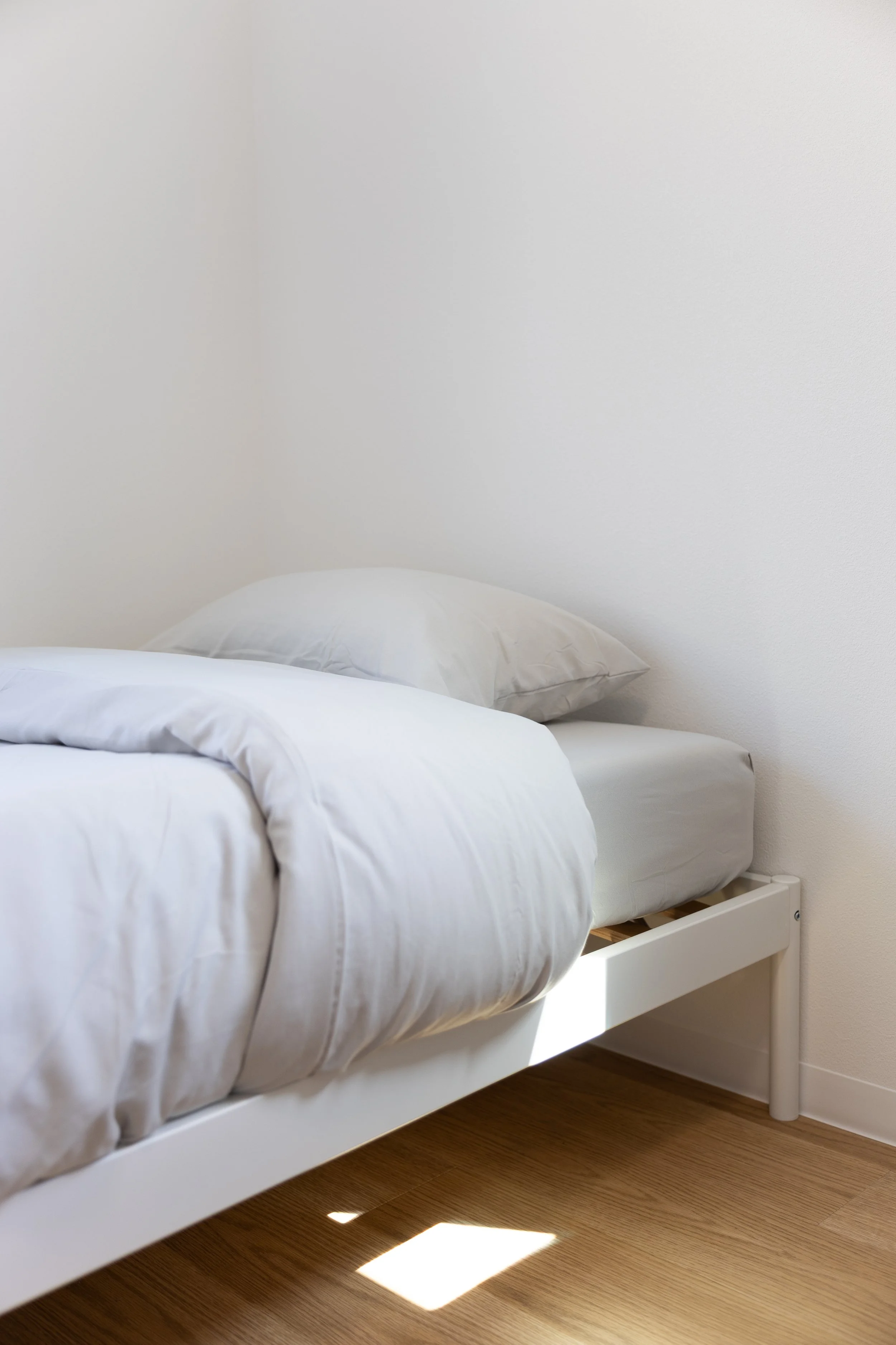 A white bed with white pillows and a white duvet cover, positioned next to a white nightstand, on a hardwood floor with sunlight coming through a window.