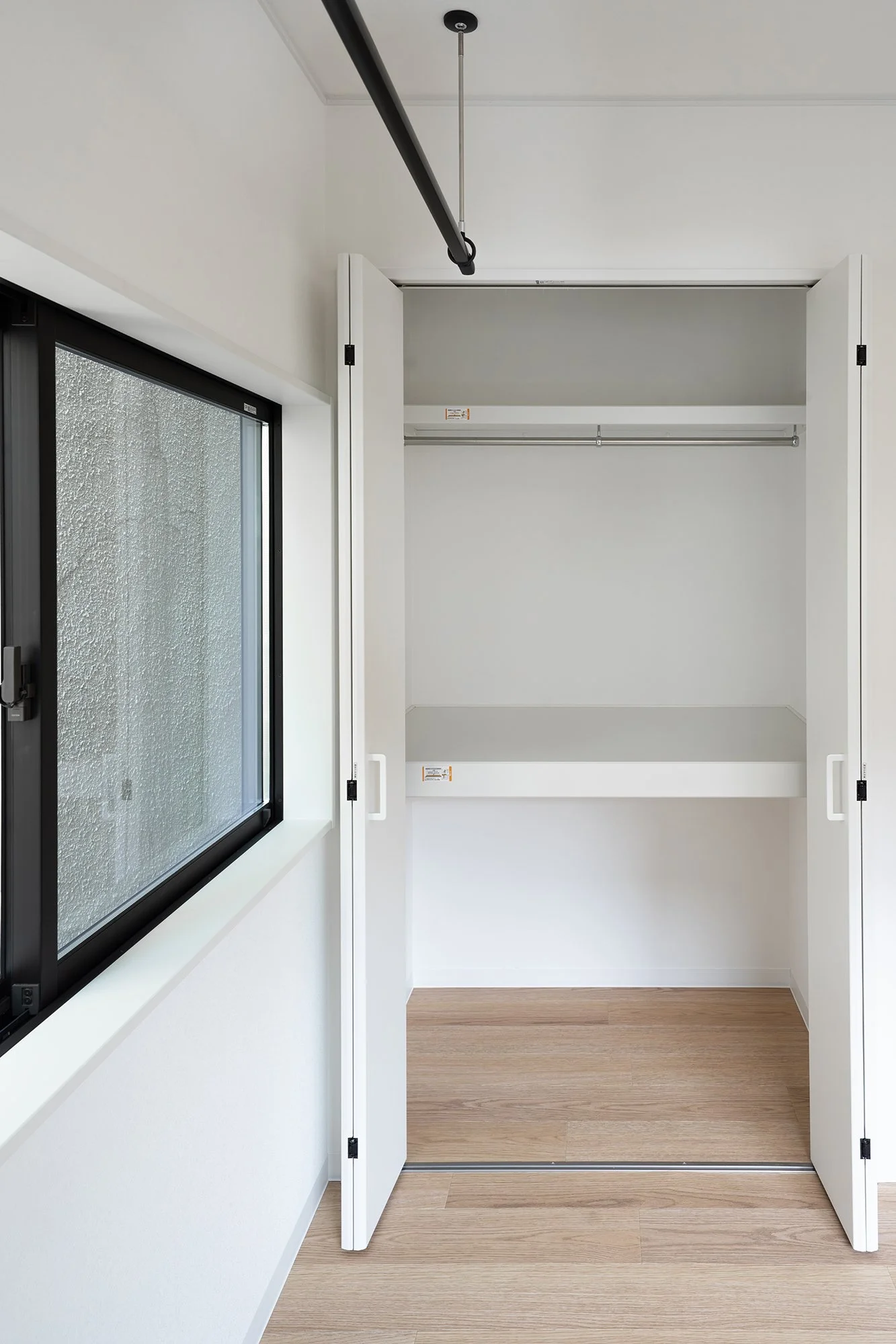 Empty white closet with open doors, a shelf, and a hanging rod. Next to a window with frosted glass and black frame.