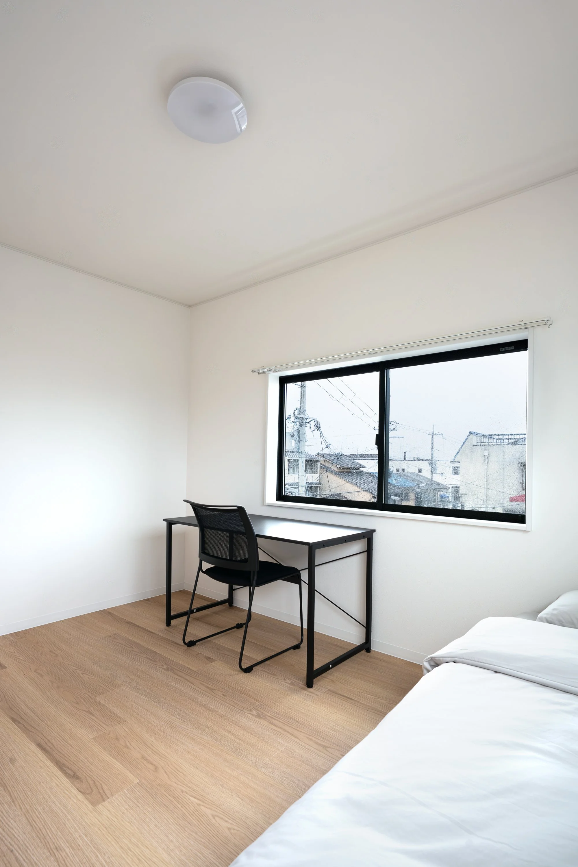 A minimalist bedroom with a bed, black desk and chair, a window with a city view, and a ceiling light