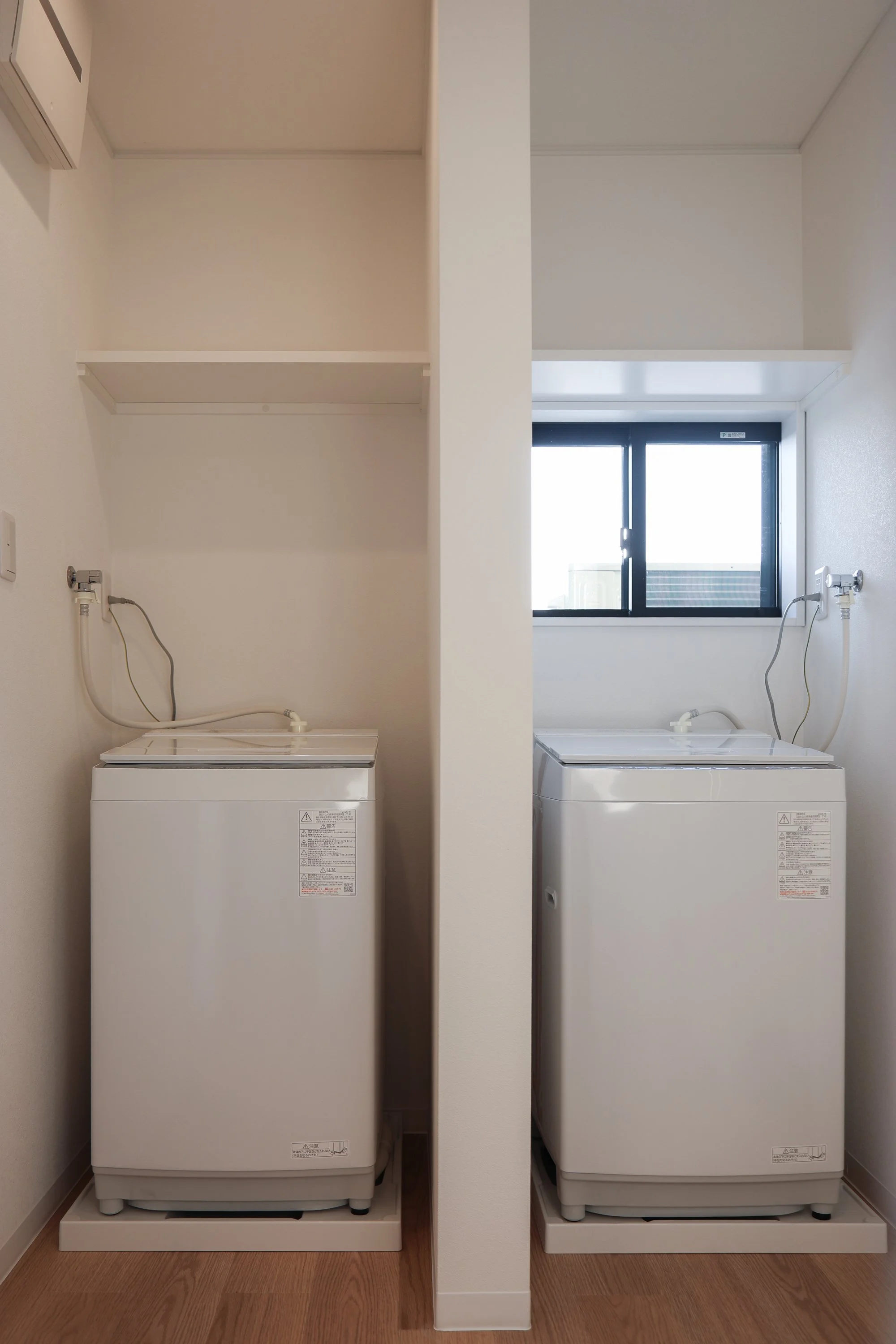 Two white top-loading washing machines in a laundry area, separated by a half wall, with a window behind the right machine.