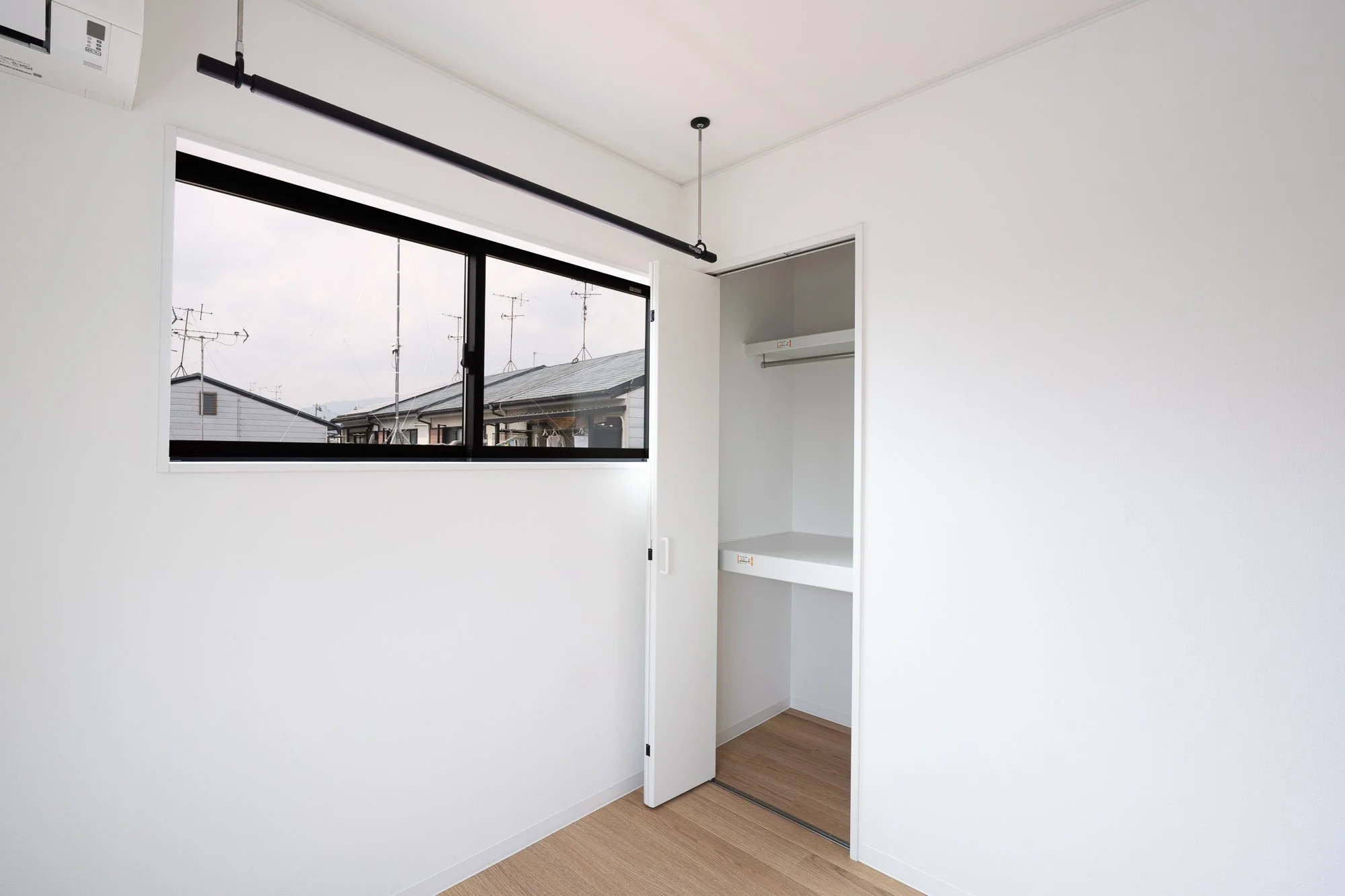 Empty bedroom with a window showing rooftops and television antennas, an open closet with a hanging rod, white walls, and wooden flooring.