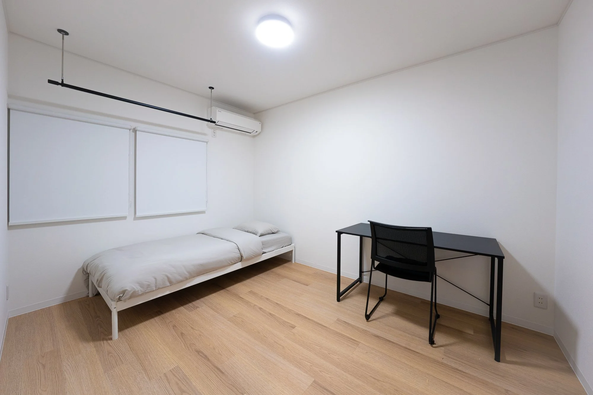 Minimalist bedroom with a single bed, black desk, black chair, white walls, light wooden floor, and air conditioning unit