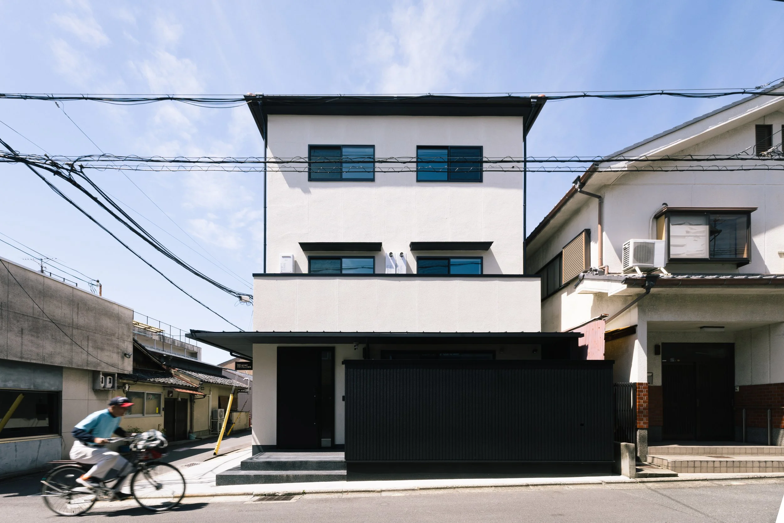 A modern white multi-story building with black window frames and a black gate, situated on a street with a person riding a bicycle in the foreground. Overhead power lines are visible against a partly cloudy blue sky.