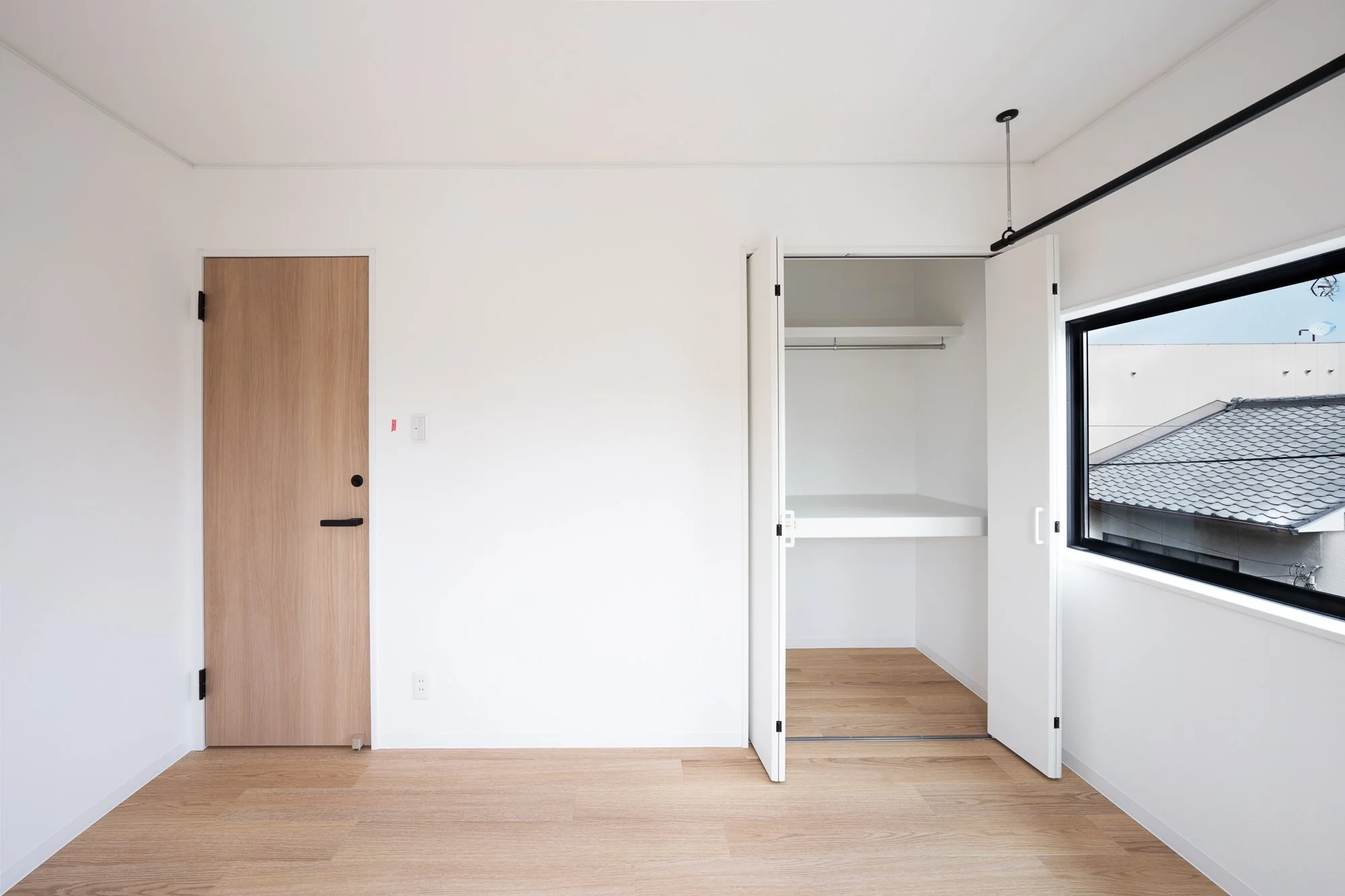 Empty room with wooden floor, white walls, a wooden door, open closet with white shelves, and a large window showing rooftops outside.