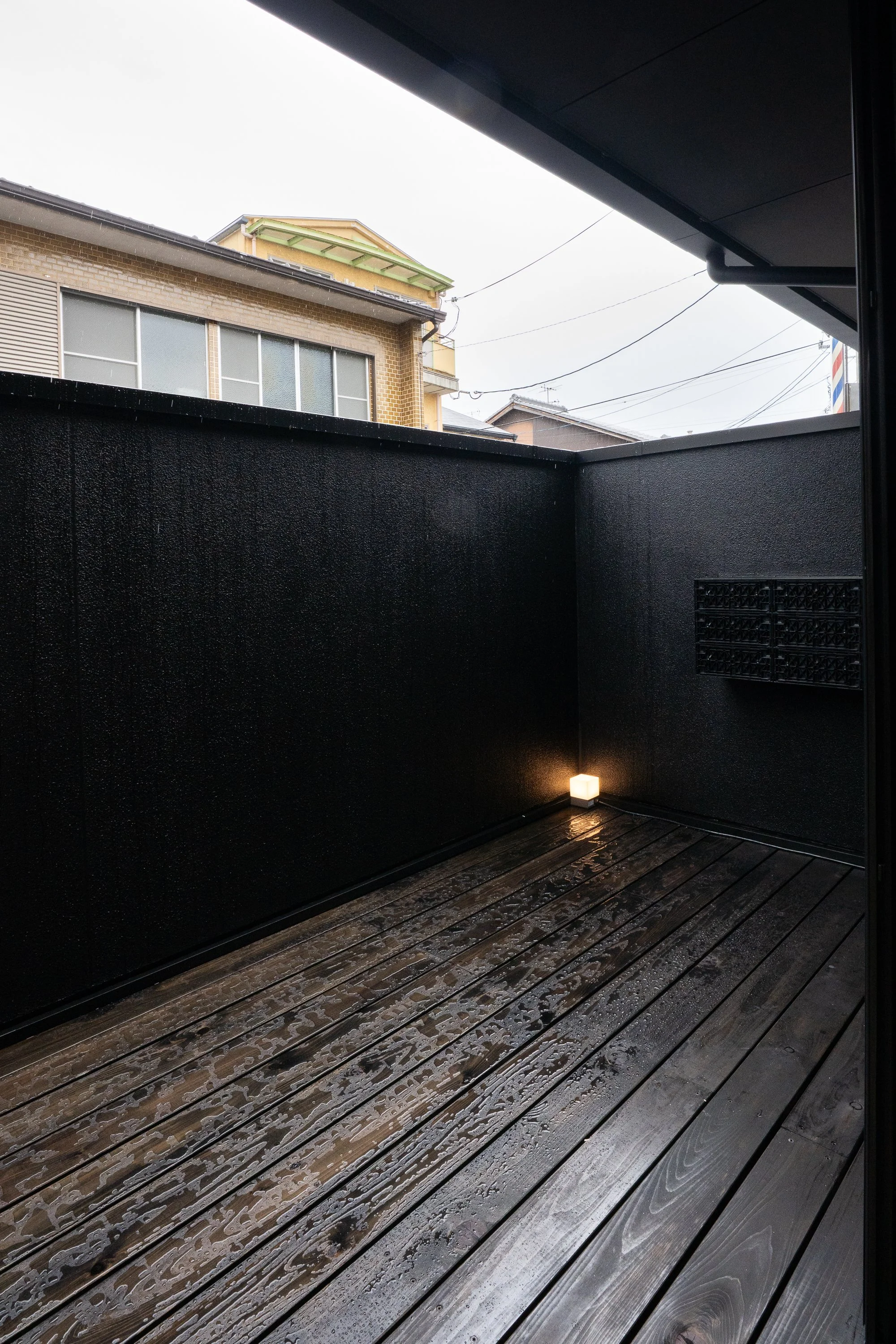 A small, empty balcony with dark walls, wet wooden floor, a small wall-mounted light, and a view of neighboring buildings and power lines in the background.