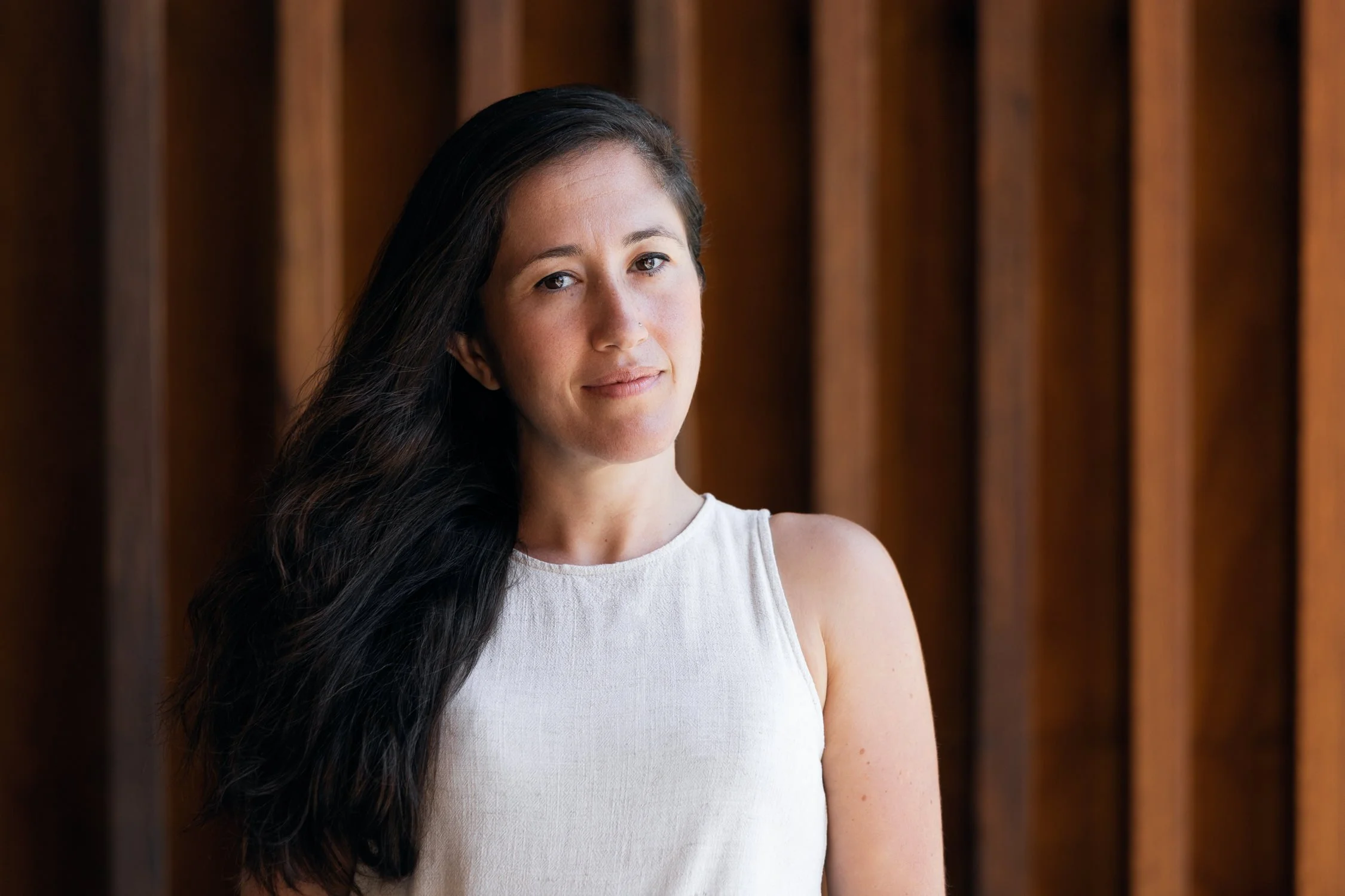 Young woman with long, dark hair wearing a white top standing in front of wooden panels.