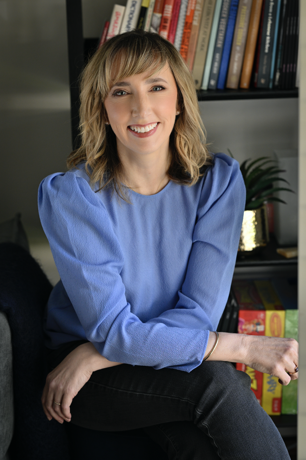 A woman with shoulder-length blonde hair and a bright smile, wearing a light blue blouse, sitting on a dark chair or sofa in front of a bookshelf with colorful books and a green plant in the background.
