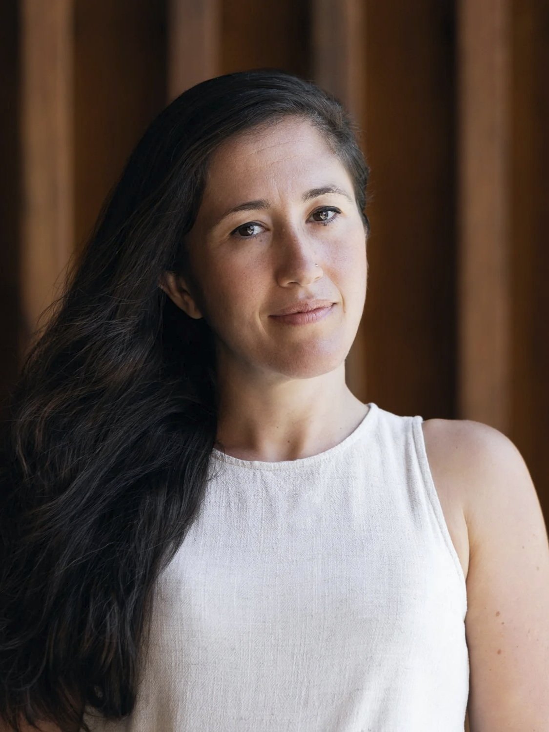 A young woman with long dark hair and light skin, wearing a sleeveless white top, standing against a wooden background.