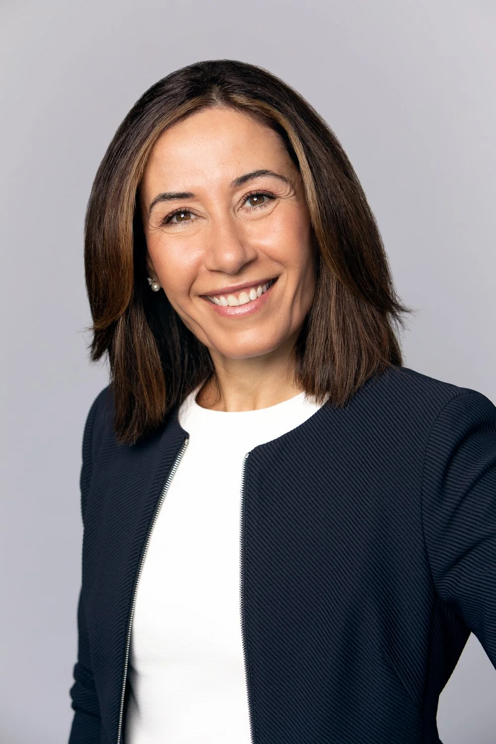 Professional woman with shoulder-length brown hair, smiling, wearing a white top and a dark navy blazer, against a light gray background.
