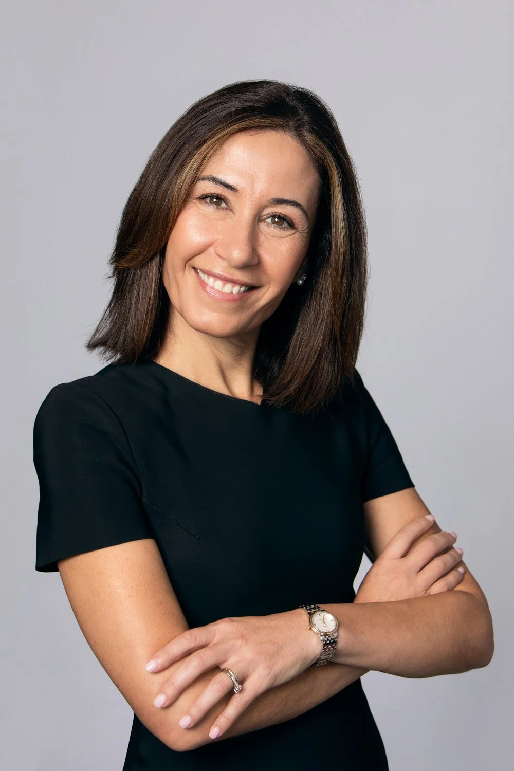 A woman smiling with arms crossed, wearing a black top and a watch on her left wrist, against a plain gray background.