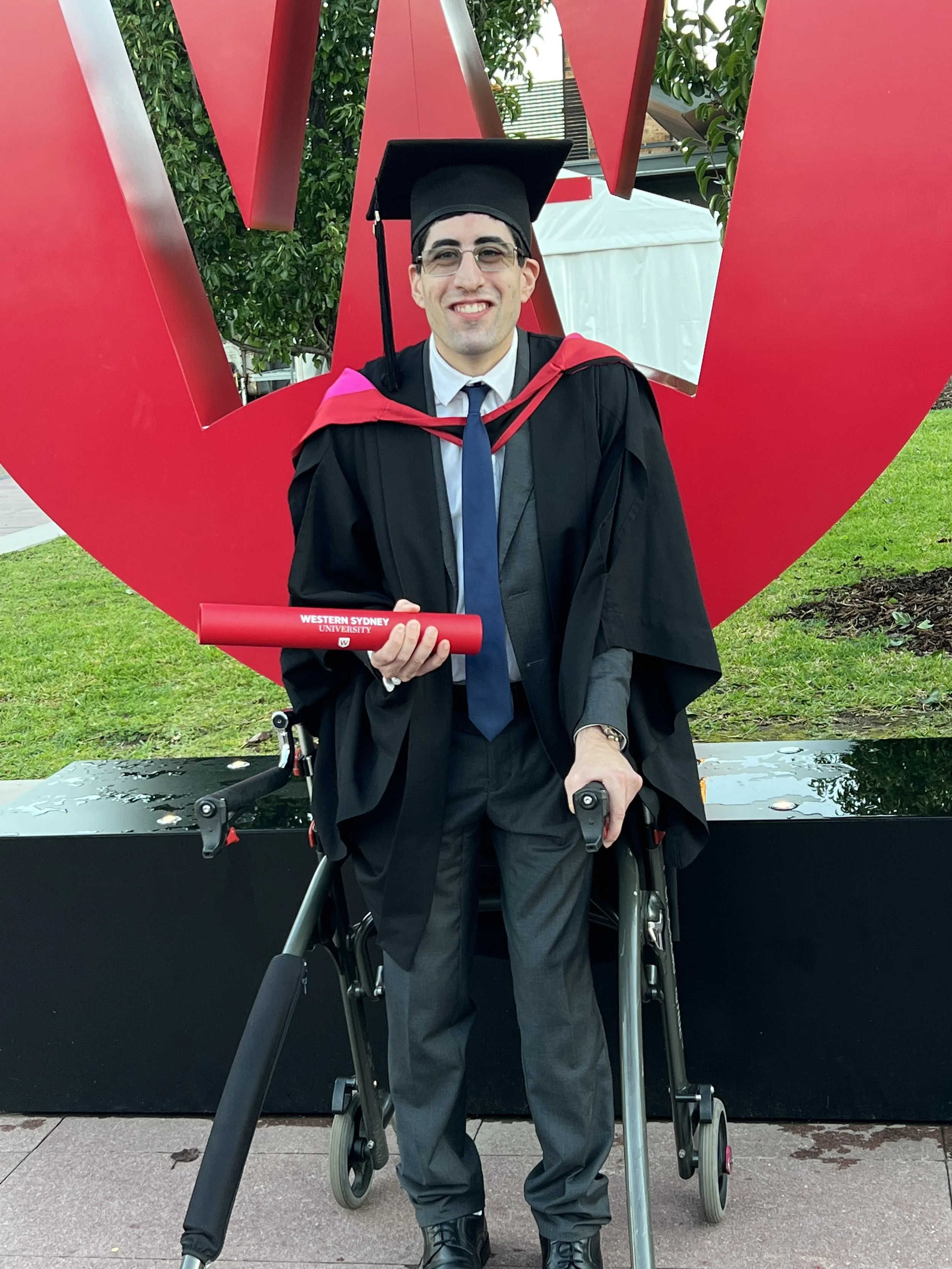 A person in a graduation gown and cap, holding a red diploma, standing in front of a large red sculpture with greenery and a building in the background.