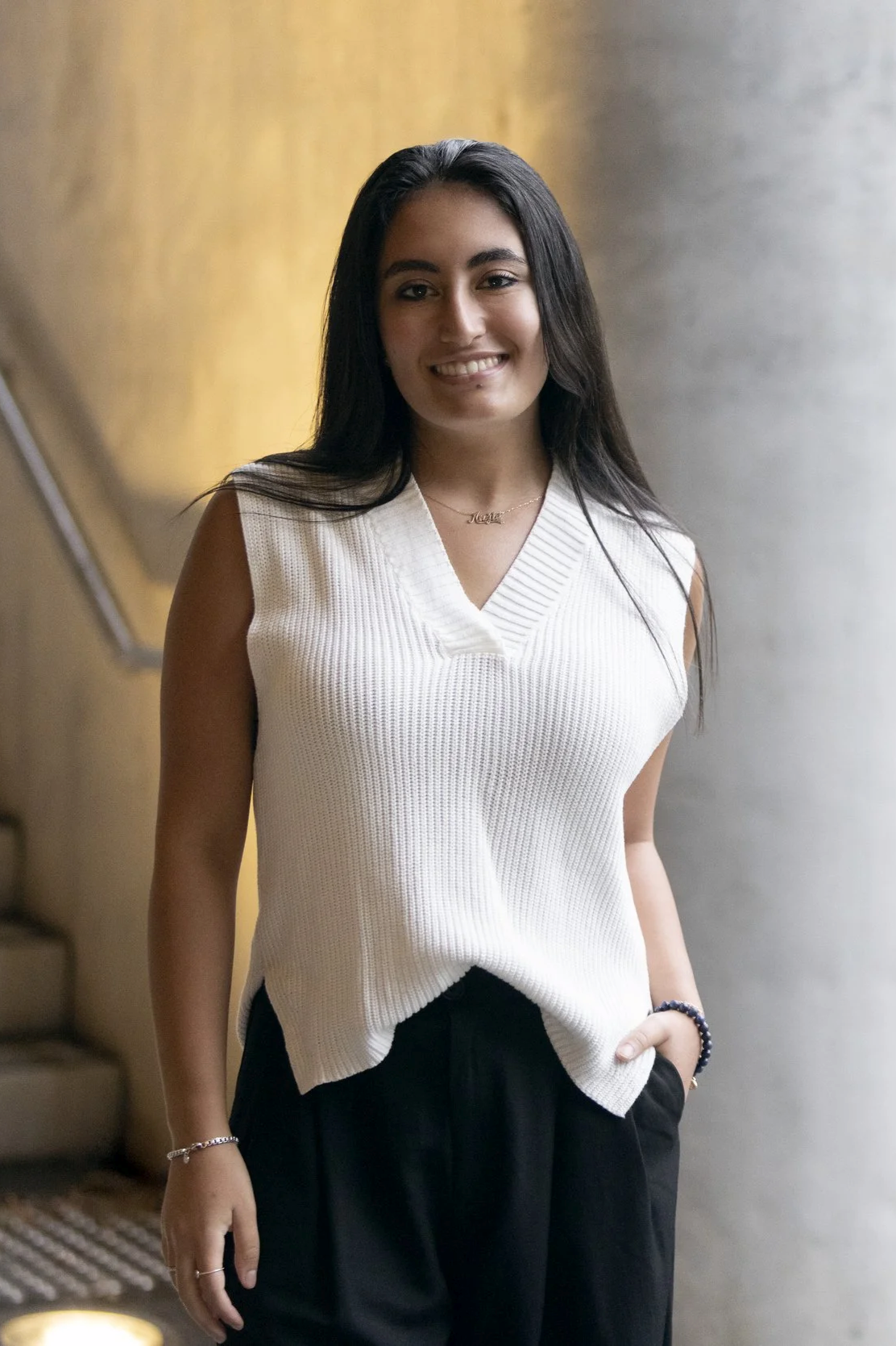 A young woman with long dark hair standing indoors, smiling, wearing a white sleeveless knitted top and black pants.
