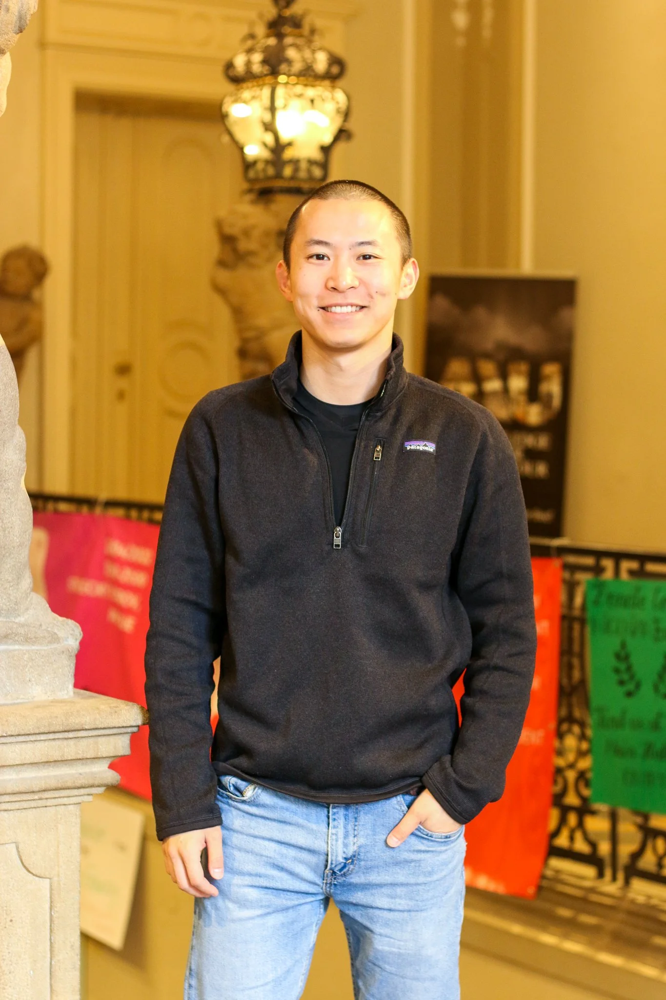 A young man with a shaved head smiling, wearing a black zip-up Patagonia fleece and light blue jeans, standing indoors with colorful bags in the background.