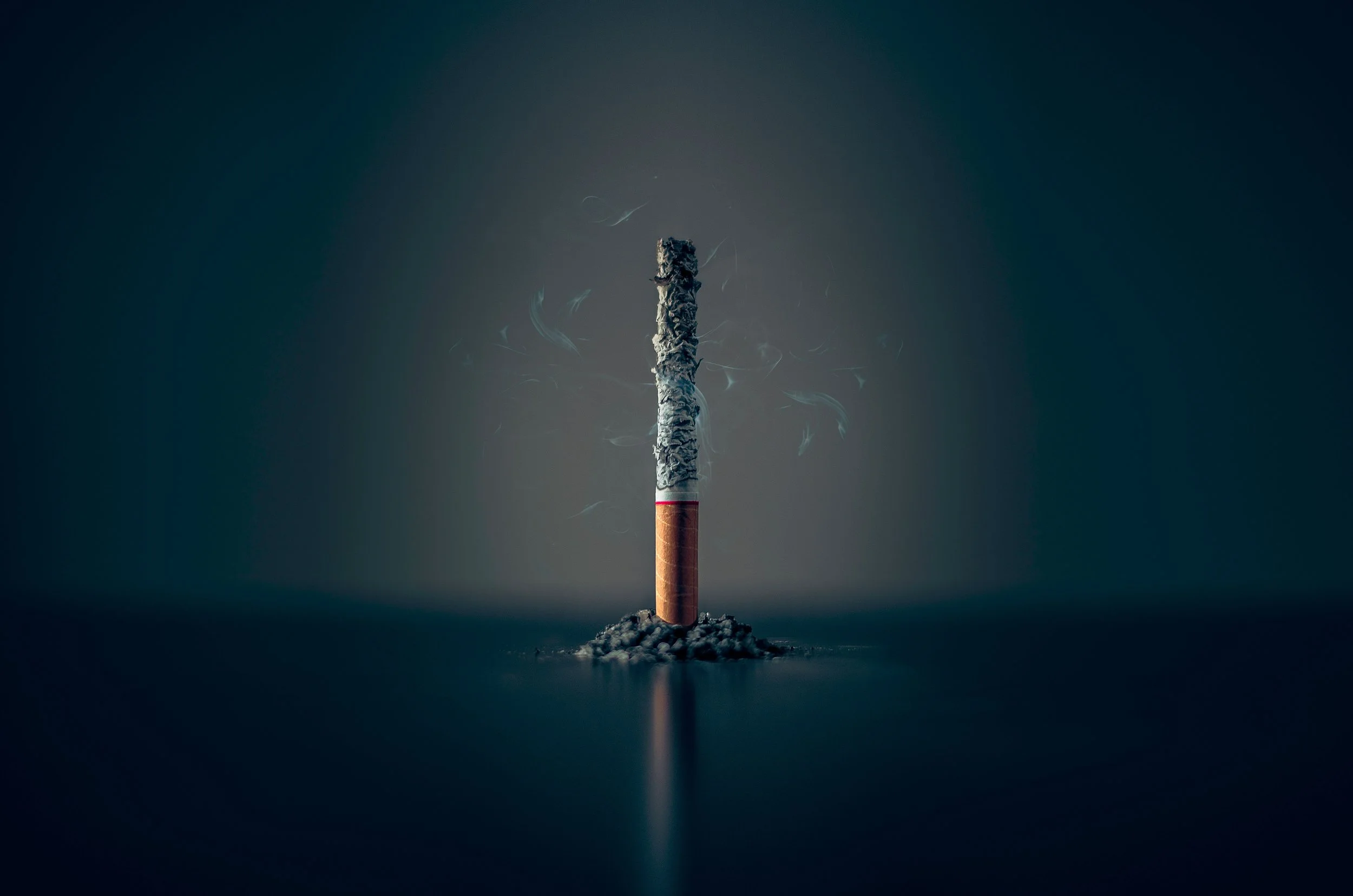 An unlit cigarette with ash at the tip is standing upright on a dark reflective surface against a plain dark background.
