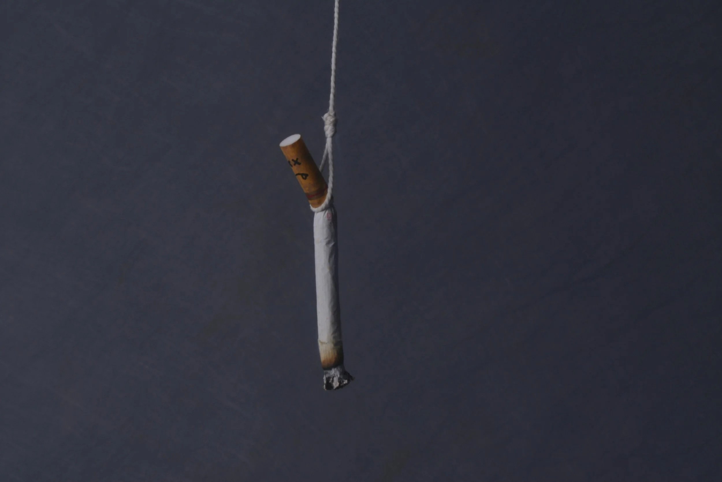 A lit cigarette with ash at the tip, hanging from a piece of string against a dark background.