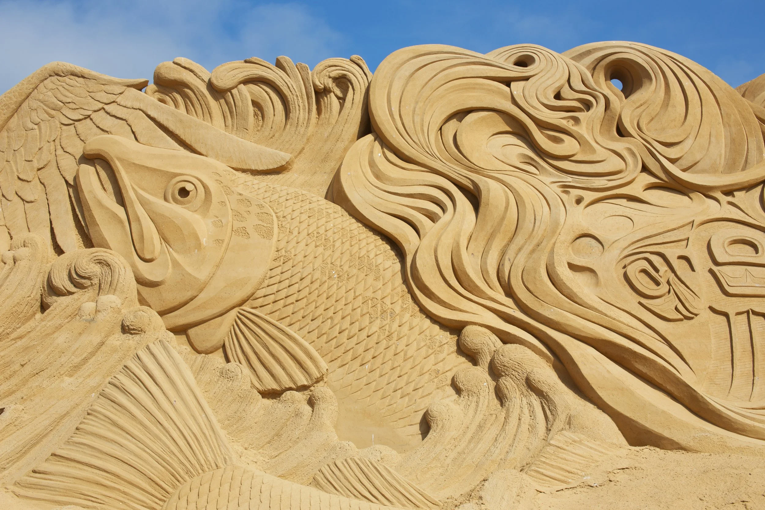 Sandsteinrelief eines mythologischen Wesens, möglicherweise ein Drache oder eine Sirene, mit detaillierten Wellen- und Schuppenmustern, vor einem blauen Himmel.