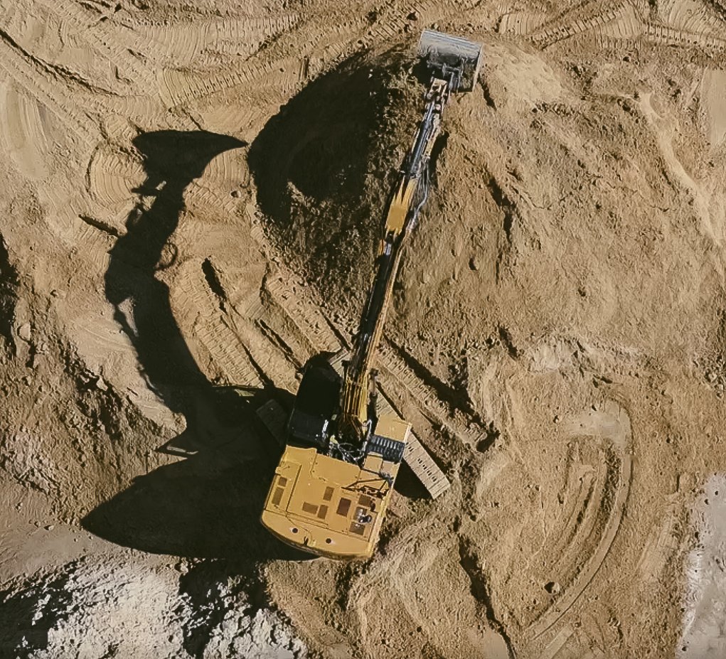 Ein Bagger arbeitet an einer Baugrube im Sand, es sind Erdbewegungsarbeiten zu sehen.