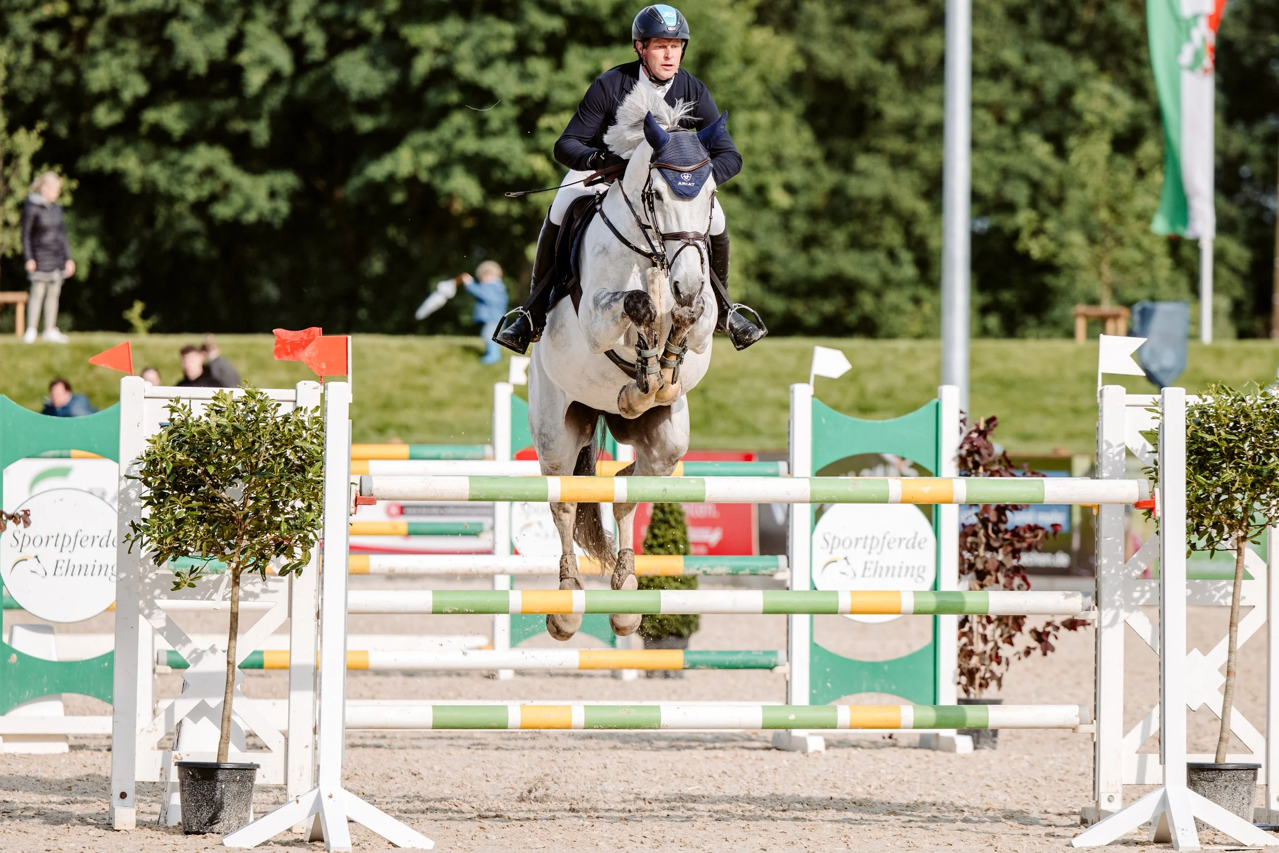 Reiter springt mit seinem Pferd über einen Spring Parcours während eines Reitwettkampfs im Freien, im Hintergrund sind Bäume und Zuschauer sichtbar.