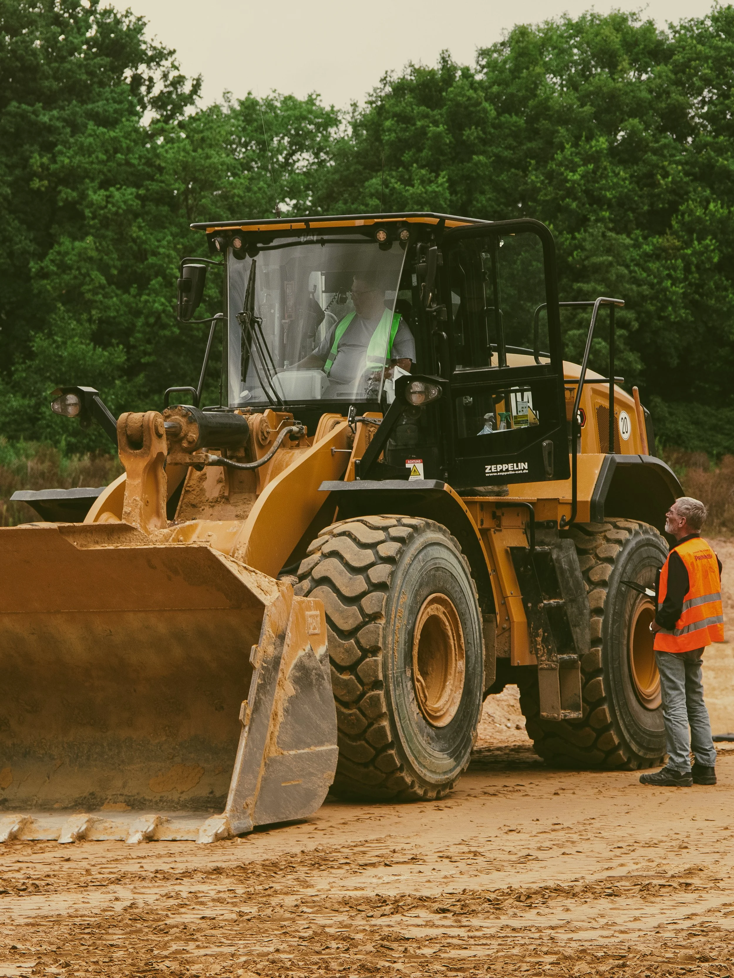Ein Bagger auf einer Baustelle, bedient von einem Fahrer in einer Kabine, während eine Person mit orangener Warnweste daneben steht und spricht, im Hintergrund grüne Bäume.