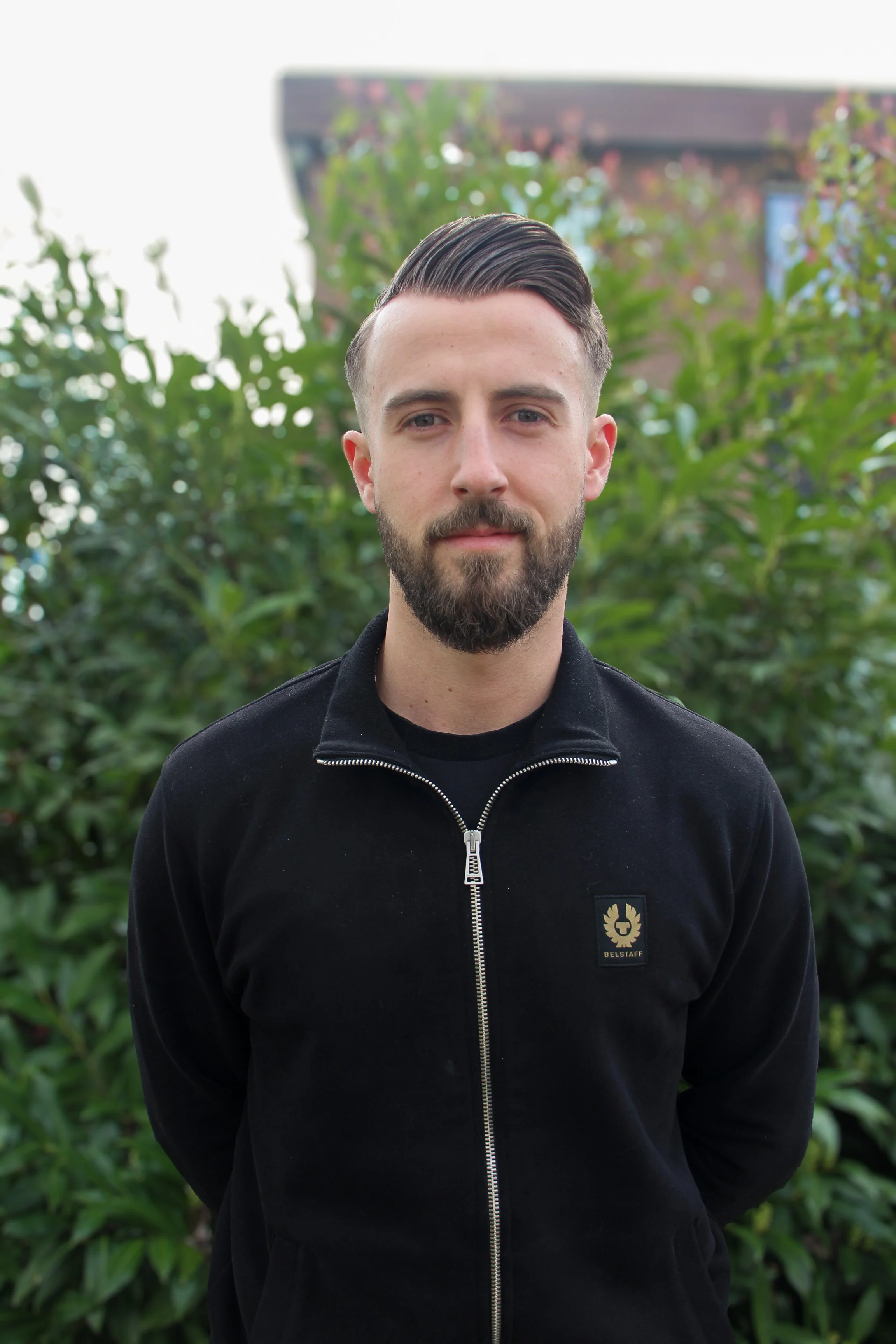Junger Mann mit dunklem Haar, Vollbart und blauen Augen, trägt eine schwarze Jacke mit Reißverschluss und Logo, steht vor einer grünen Hecke im Freien.