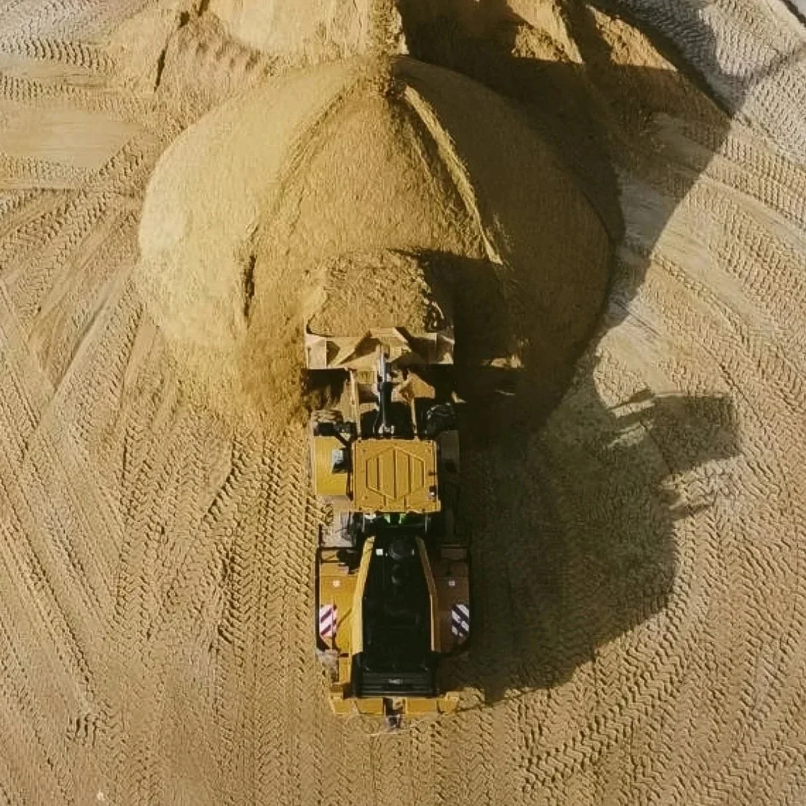 Ein Baugerät, das auf einer Sandfläche arbeitet, wahrscheinlich ein Radlader, mit sichtbaren großen Rädern und einer Schaufel, die vorwärts bewegt wird.