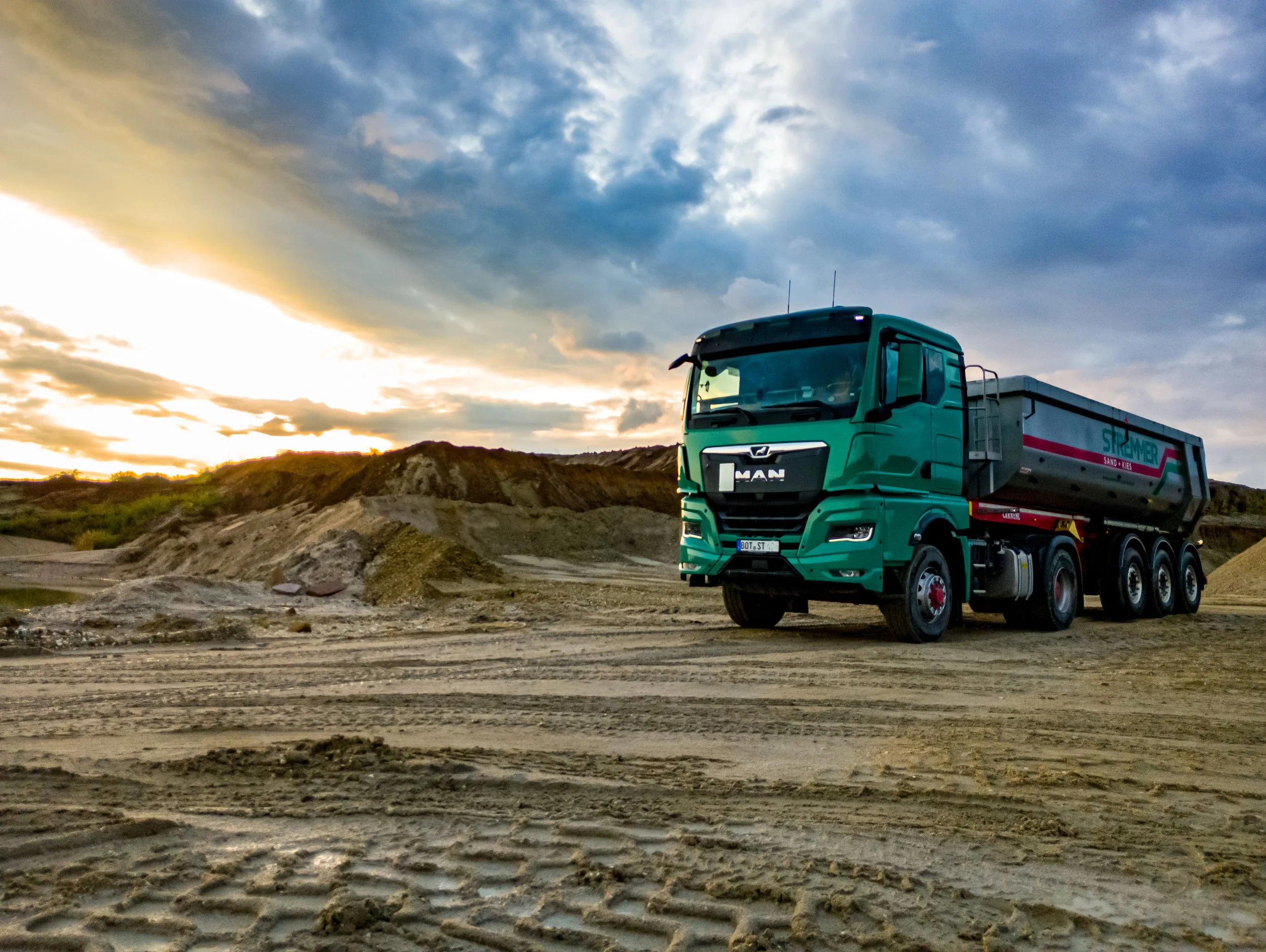 Ein grüner MAN-LKW fährt durch eine unbefestigte, sandige Landschaft bei Sonnenuntergang, mit bewölktem Himmel im Hintergrund.