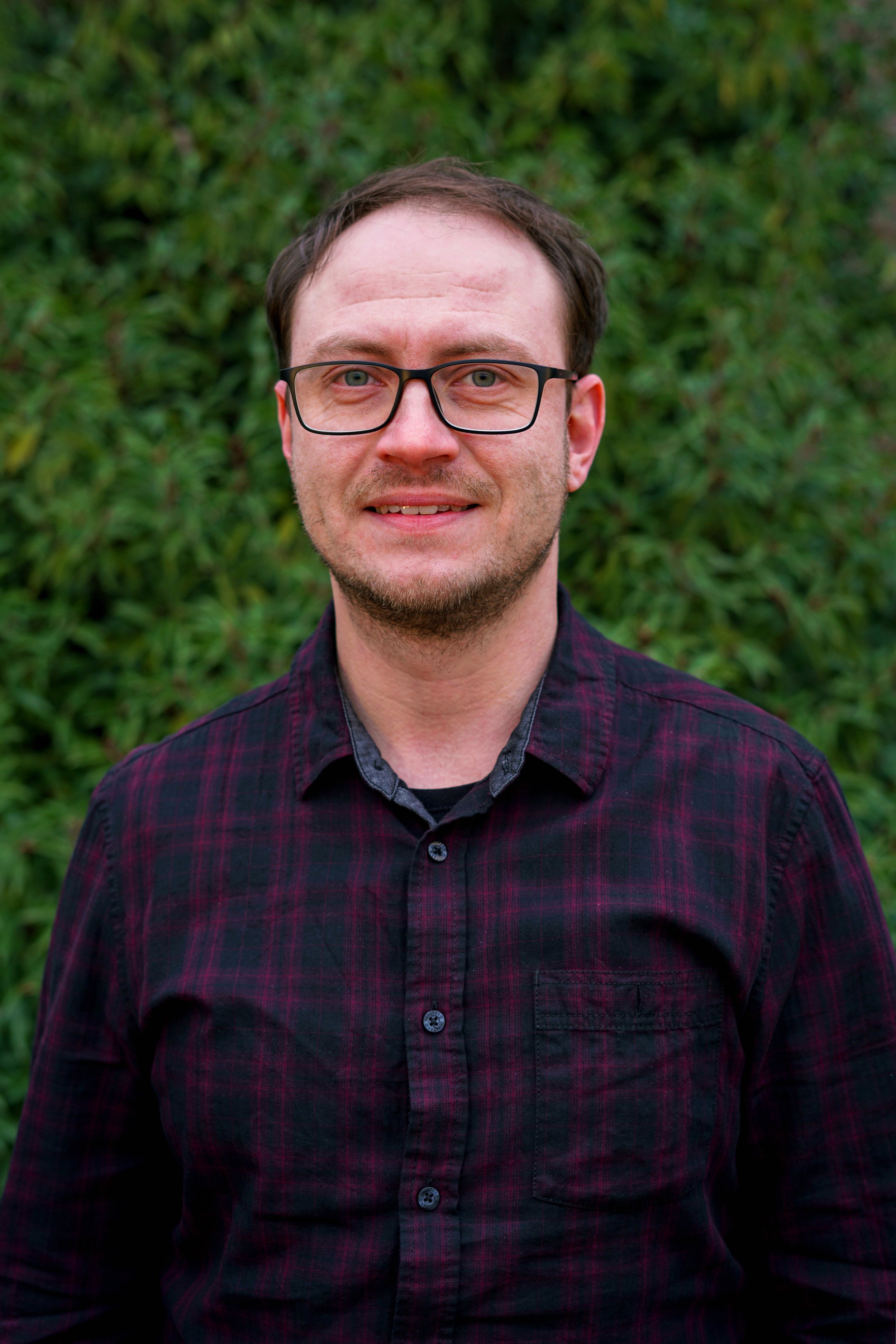 Porträt eines Mannes mit dunklen Haaren, Brille und braunem Bart, trägt ein dunkelrotes kariertes Hemd, vor grünem Büschen-Hintergrund.