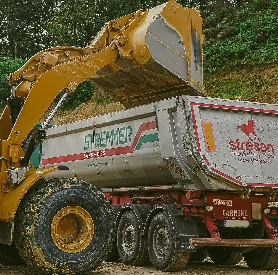 Ein Baustellenfahrzeug mit gelbem Bagger, der Sand und Kies in einen Muldenwagen lädt. Das Fahrzeug trägt Labels mit der Aufschrift 'Stremmer', 'Stresan' und 'Carnehl'.